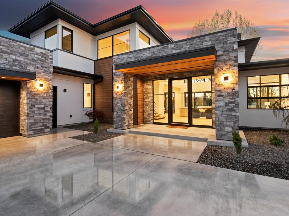 Eagle, Idaho - Eco Builders Modern house exterior with large windows, stone facade, and a concrete driveway at sunset.