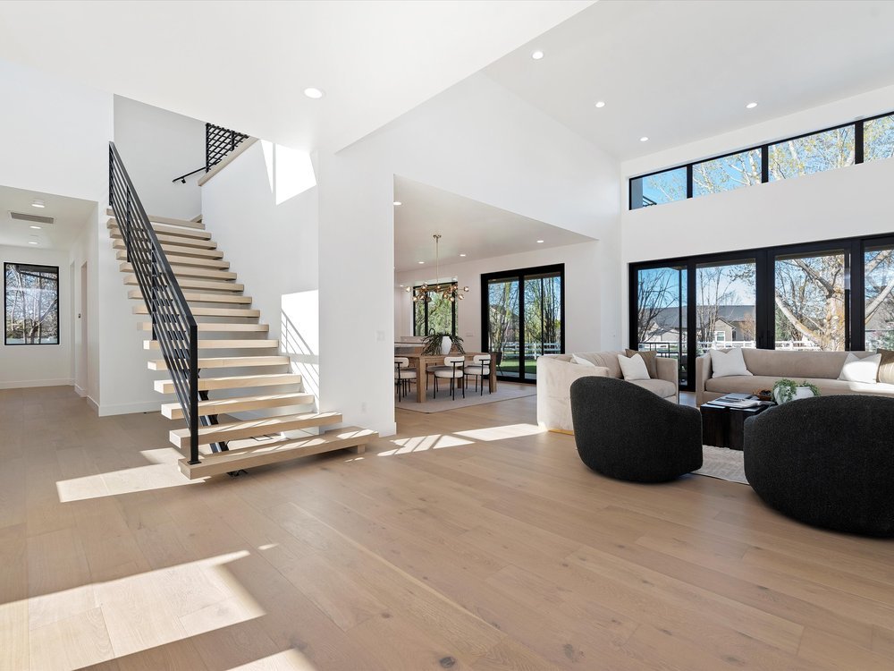 Eagle, Idaho - Eco Builders Modern open-concept living space with a floating staircase, large windows, sectional sofa, and round black chairs.