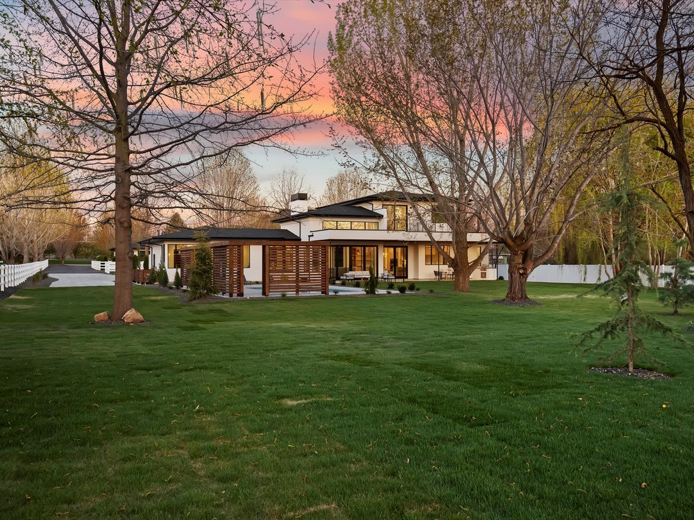 Eagle, Idaho - Eco Builders Modern two-story house with large windows, surrounded by a green lawn and trees, under a sunset sky.