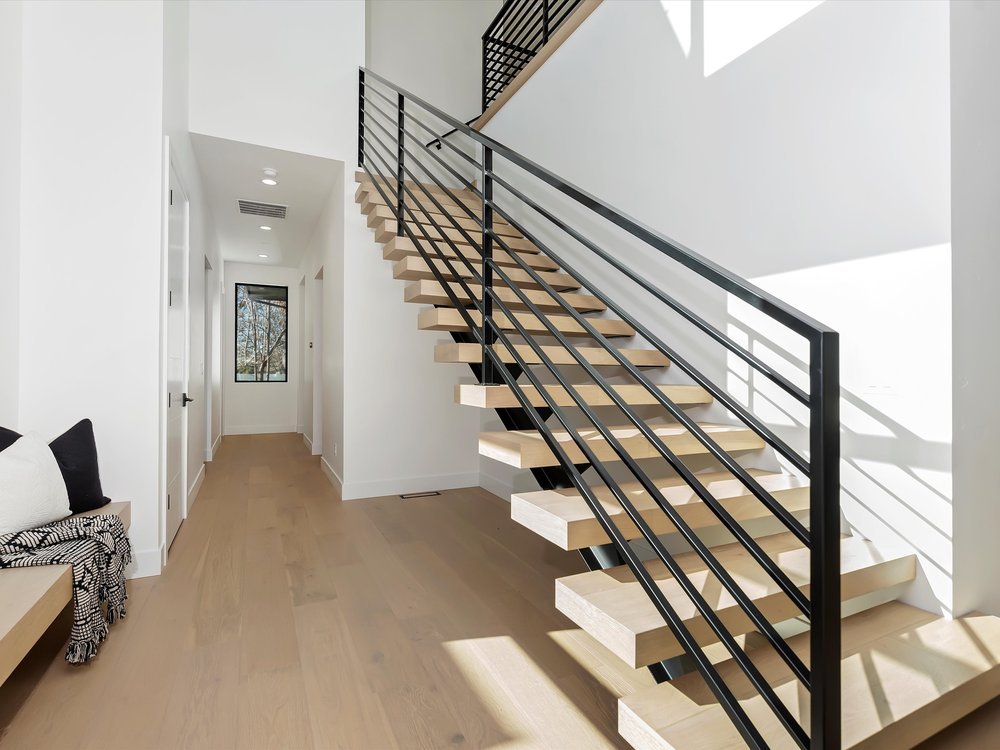 Eagle, Idaho - Eco Builders Modern interior with wooden staircase and metal railing, white walls, hardwood floor, and a hallway leading to a window.