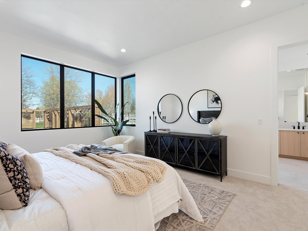 Eagle, Idaho - Eco Builders Modern bedroom with large windows, a white bed, beige knit throw, decorative pillows, a black dresser with circular mirrors above, and a potted plant in the corner.