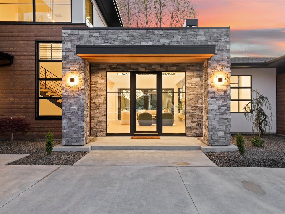 Eagle, Idaho - Eco Builders Modern house entrance with stone and wood facade, large glass doors, exterior lighting, and a landscaped front yard during sunset.