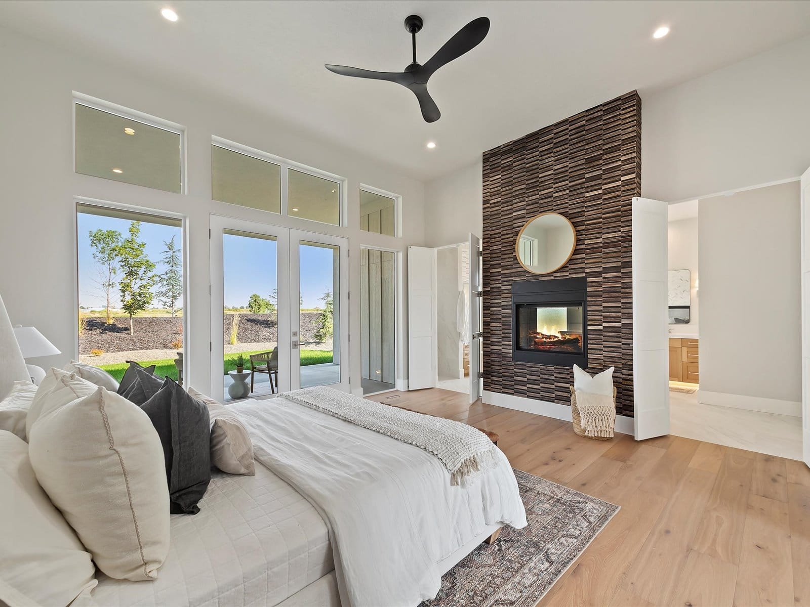 Eagle, Idaho Homes, LLC - Modern bedroom with large windows and garden view, featuring a bed with white linens, decorative pillows, a textured fireplace with a round mirror above, ceiling fan, and access to a bathroom.
