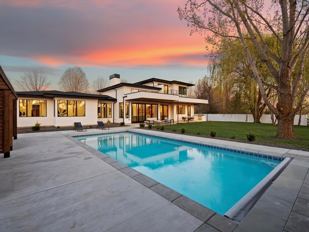Eagle, Idaho - Eco Builders Modern house with a large swimming pool in the backyard, surrounded by a patio and trees, under a colorful sunset sky.