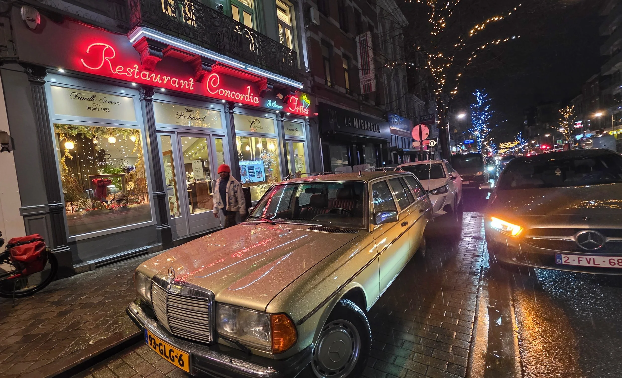 Exterior image of the Restaurant Concordia in Liege, Belgium with Christmas lights illuminating the street and a gold 1980s Mercedes estate parked in front.