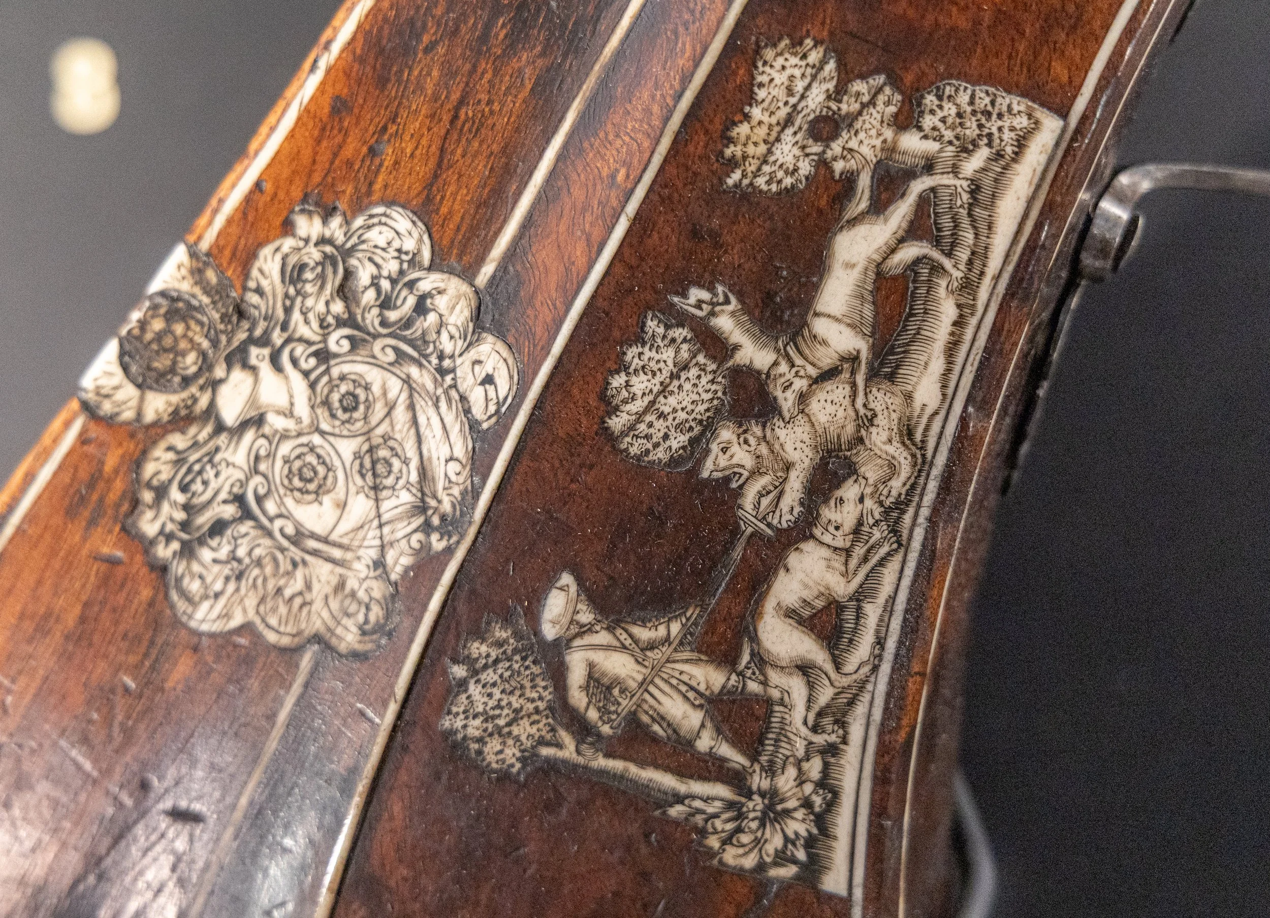 An ivory inlay on a gun stock showing a hunter spearing a bear as his two dogs have it held at bay.