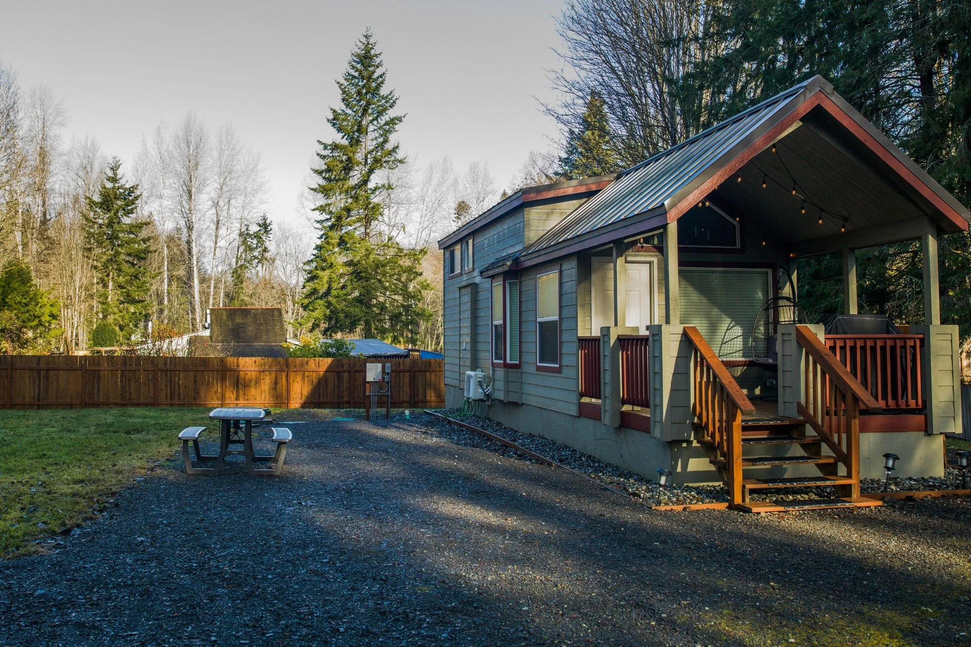 Park model RV tiny home with a covered porch on a gravel pad on private property, surrounded by evergreen trees