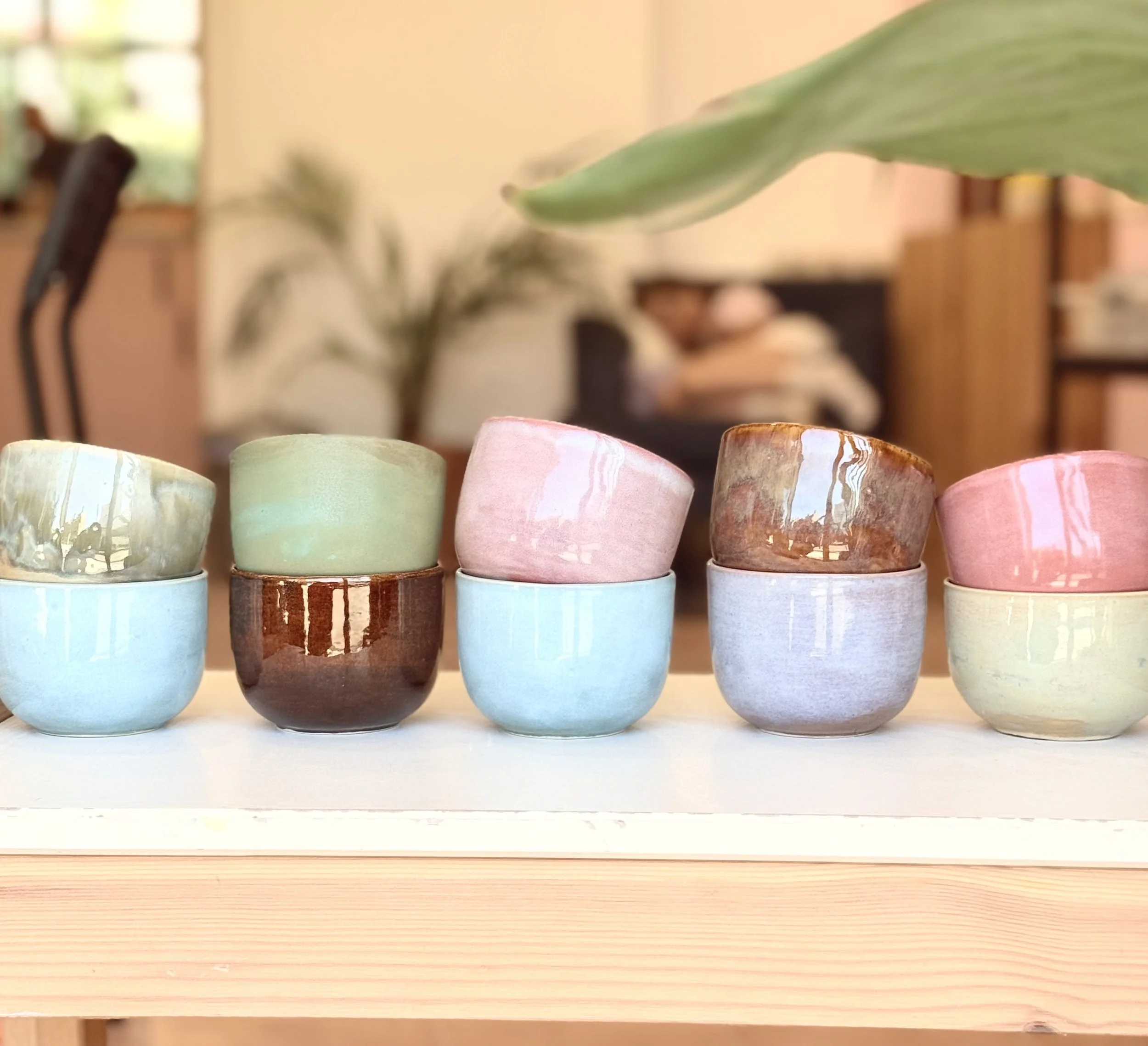 Seis tazas de cerámica en diferentes colores y acabados, apiladas en parejo en una mesa de madera, con fondo difuso de interior con plantas y muebles.