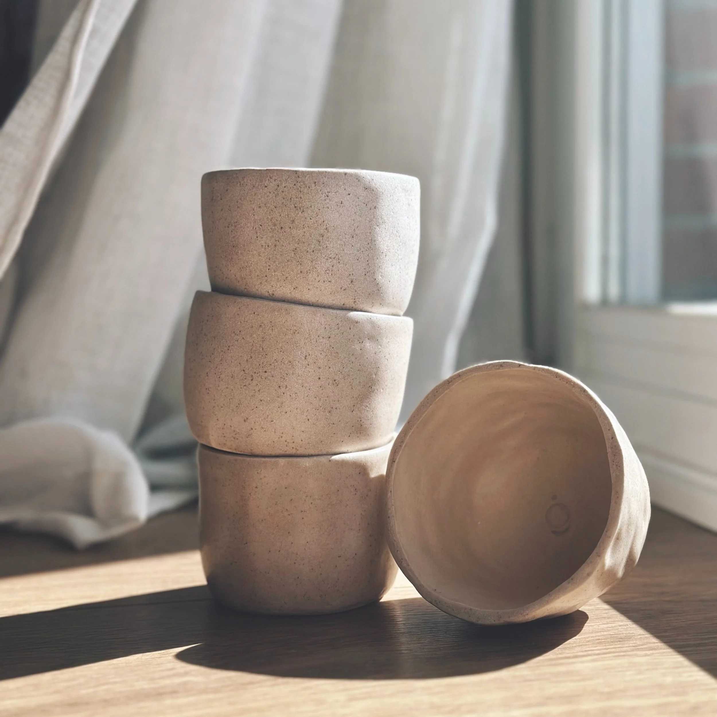 Cuatro vasos de cerámica en una superficie de madera, uno de ellos de lado mostrando su interior, en un fondo de cortinas y ventana