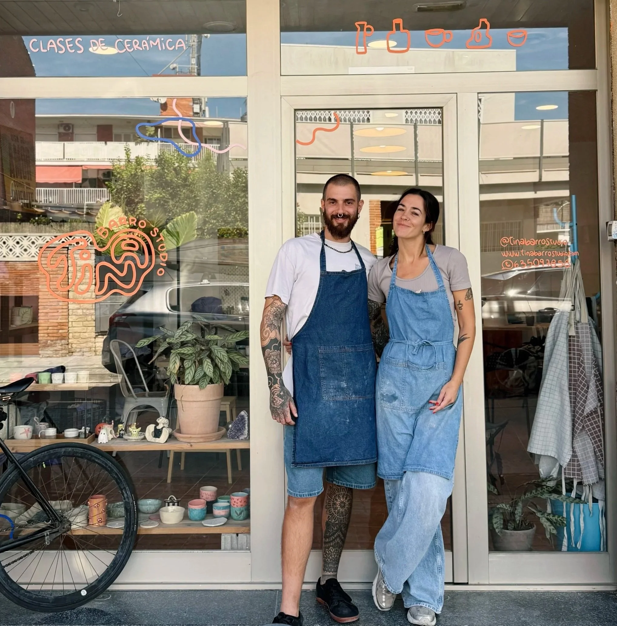 Dos personas en ropa de cocina posando frente a la entrada de un estudio de cerámica, con indicadores de cursos y decoración en la ventana.