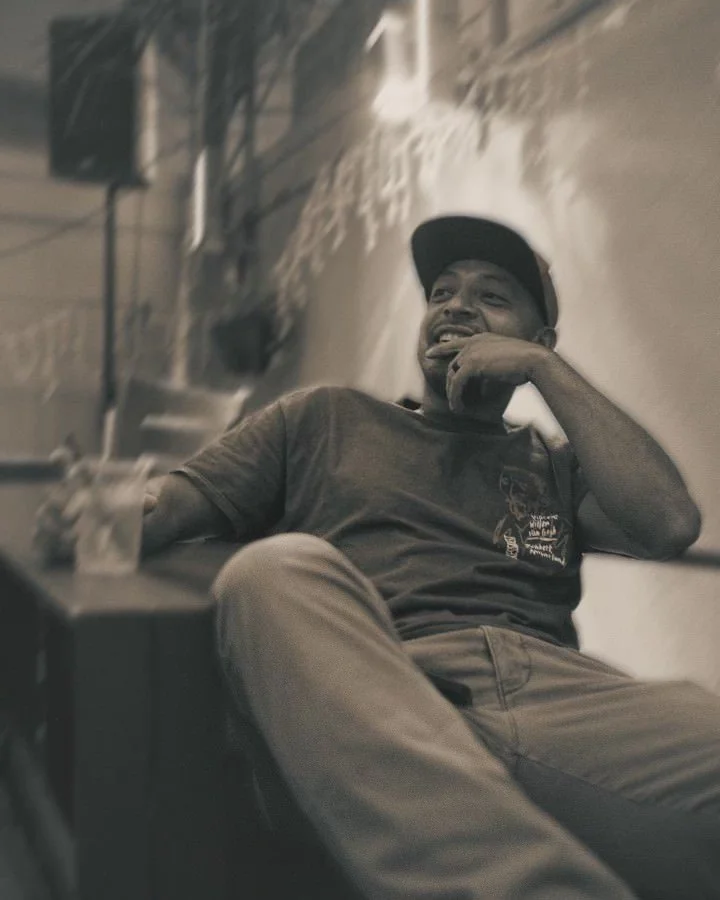 A man sitting casually at a table, smiling and laughing, wearing a baseball cap and casual clothing, with a drink and a phone on the table, in a dimly lit indoor setting.