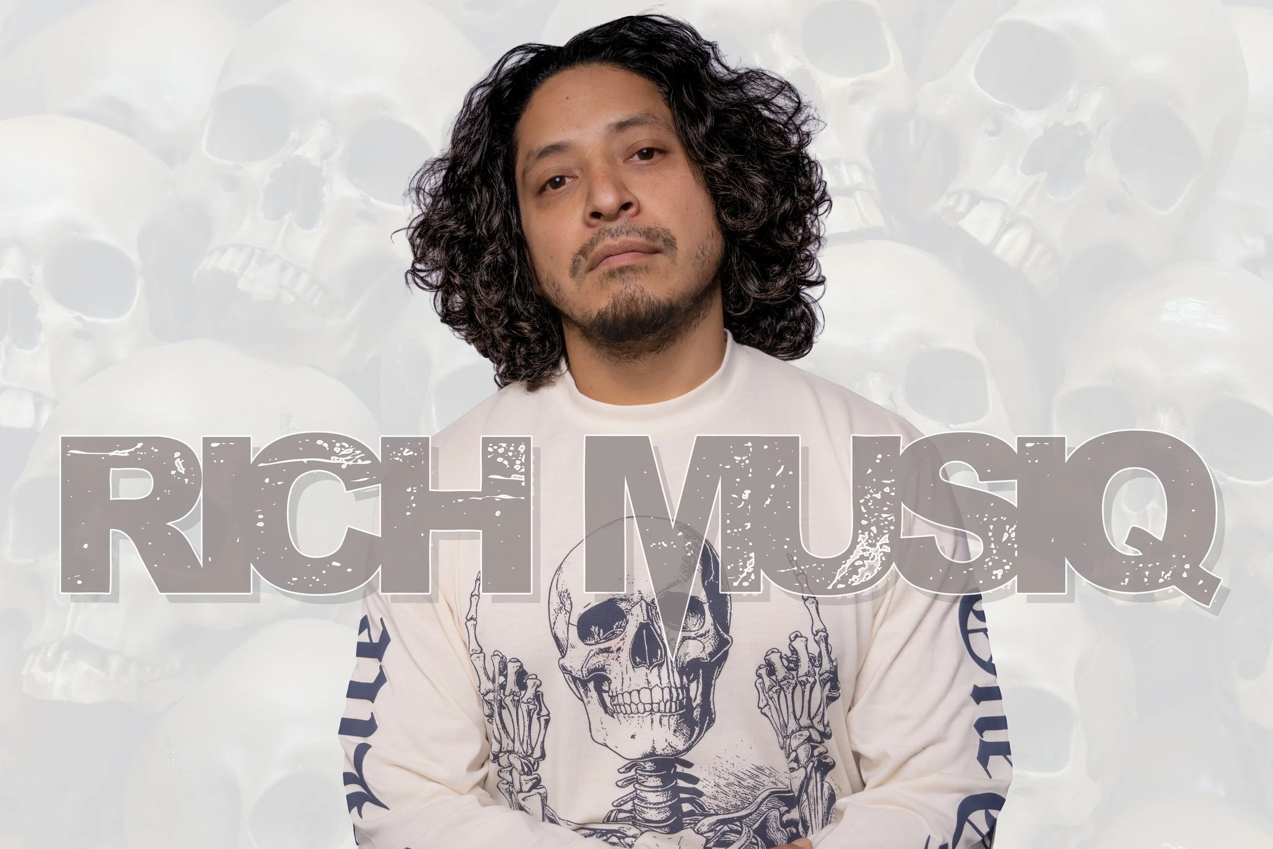 A man with long curly black hair, a goatee, and a mustache wearing a white shirt with skull print and letters, standing in front of a background with skull designs and a large text overlay.