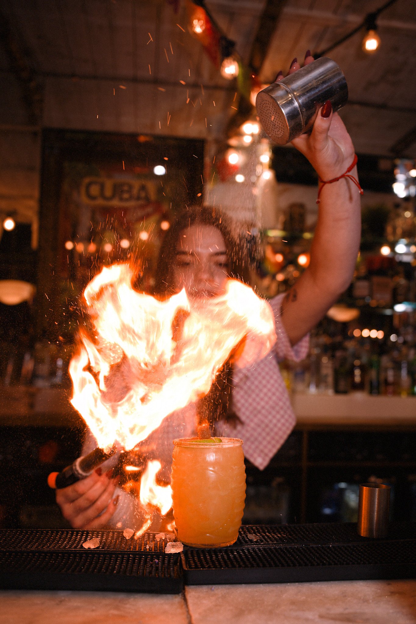 A woman sprinkles flaming alcohol into a jar, creating large flames at a bar.