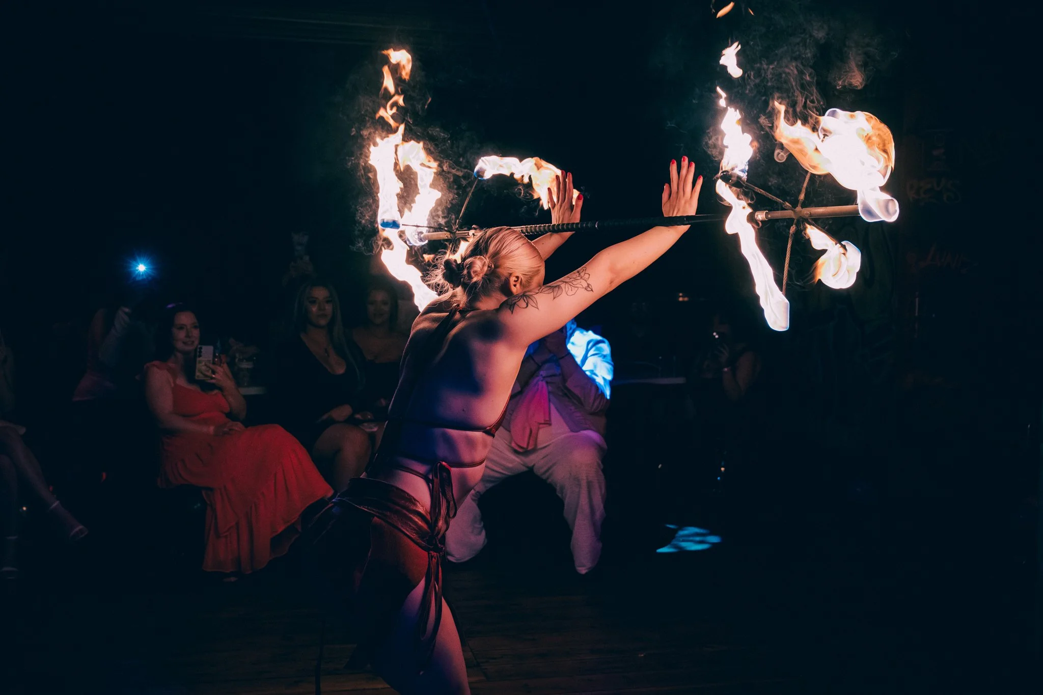 A woman with blonde hair in a bun performs a fire dancing act, holding fire poi with flames ablaze, in front of an audience in a dark venue.