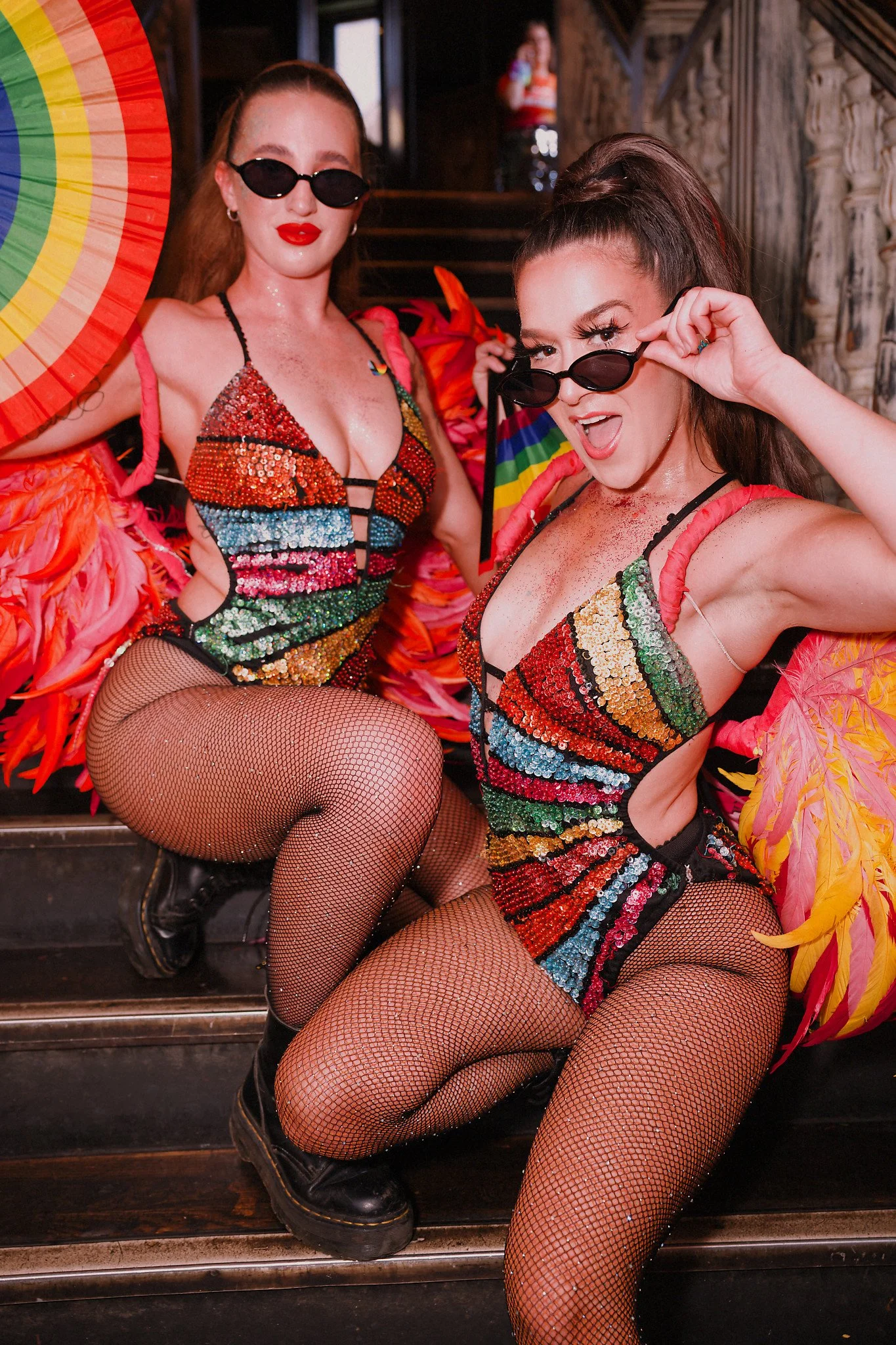 Two women with long hair wearing rainbow sequin dresses, fishnet tights, and sunglasses, sitting on stairs with rainbow decorations around them at a festive event.
