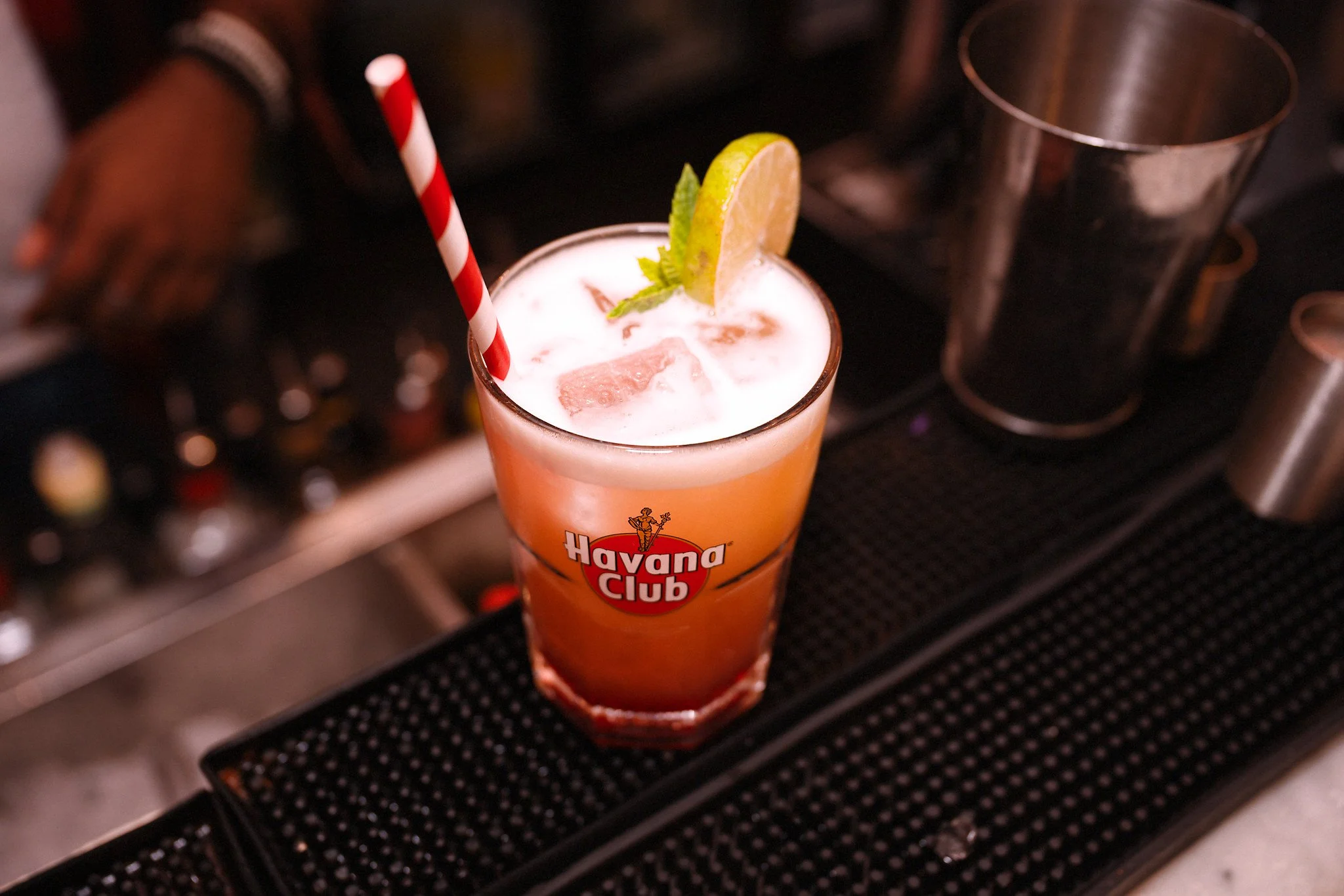 A cocktail with lime and mint garnish in a glass with a red and white striped straw, placed on a bar counter with alcohol bottles in the blurred background.