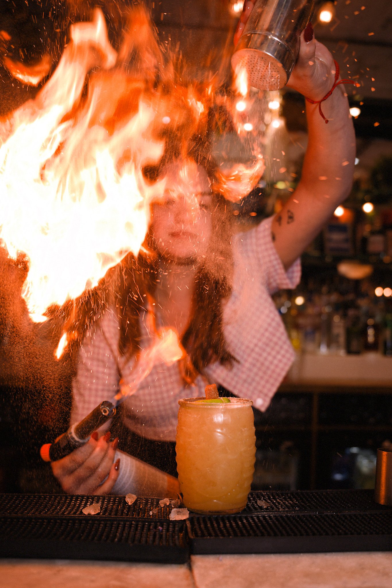 A person pouring flaming alcohol into a yellow cocktail glass on a bar counter while holding a metal shaker, with a blurred background.