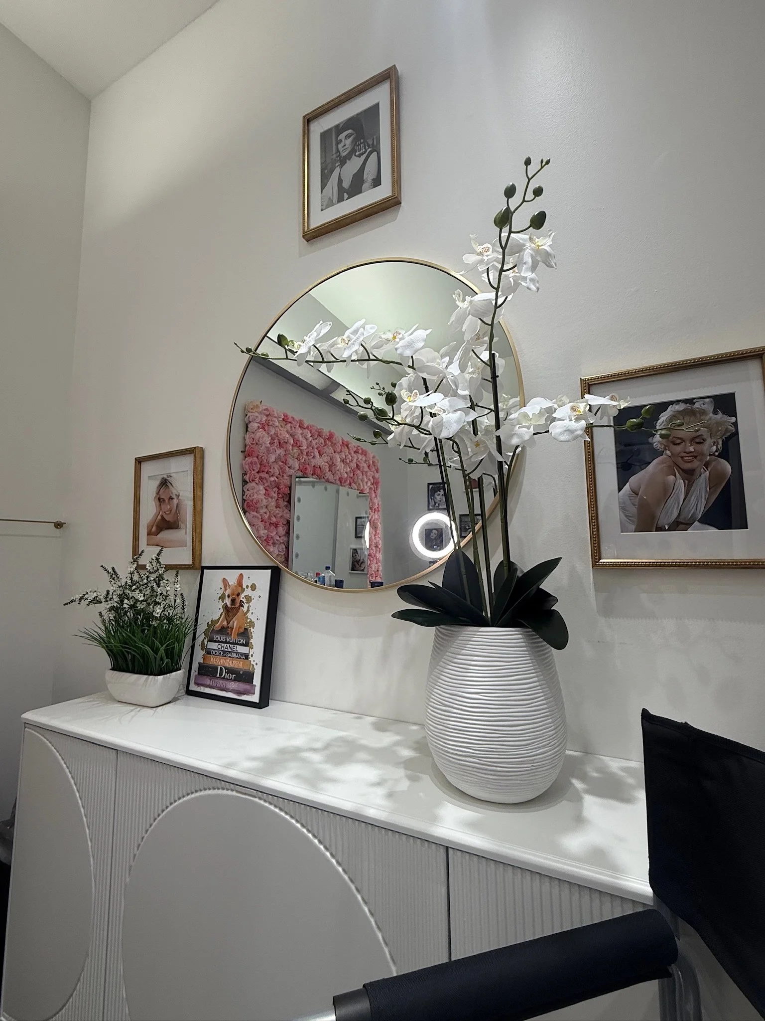 A white sideboard with decorative plants and framed photos, a round mirror reflecting a pink floral wall, and framed pictures of women on the wall.