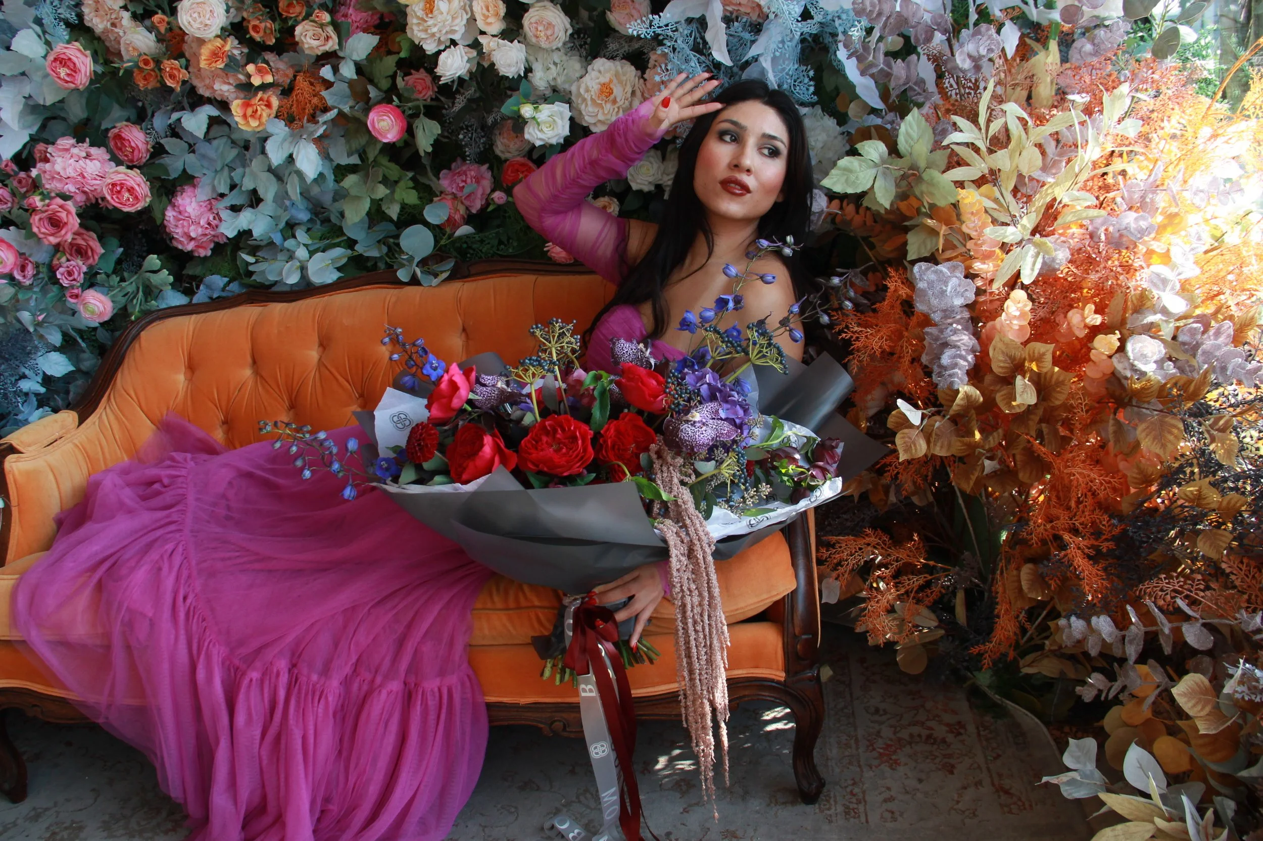 A woman in a pink dress and purple sheer sleeves reclining on an orange vintage sofa amidst a lush display of pink, white, and purple flowers with greenery and orange foliage around her.