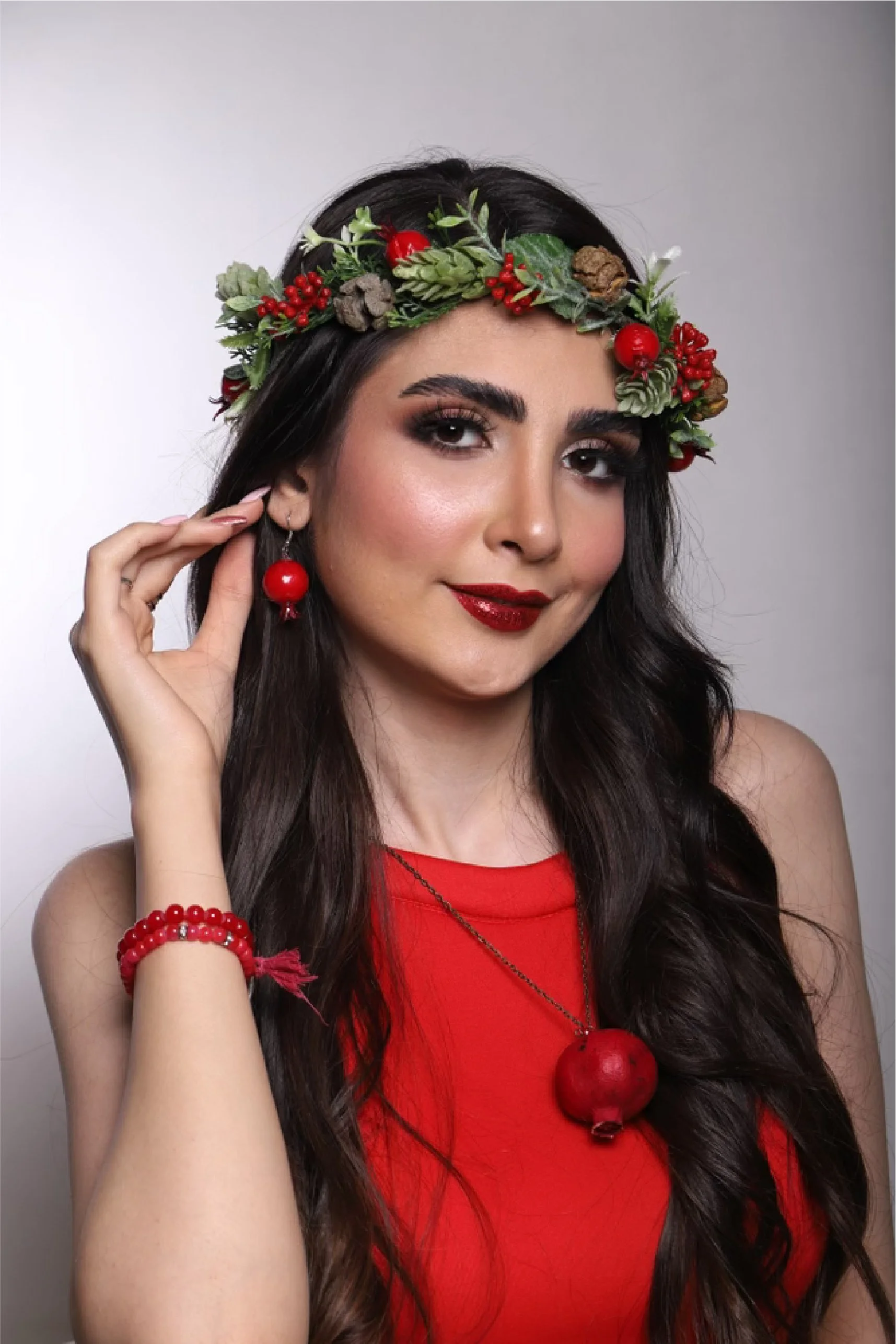 Young woman with dark hair, wearing red lipstick, red dress, and jewelry, has a festive crown made of berries, greenery, pinecones, and red ornaments, with matching earrings and a pomegranate necklace.