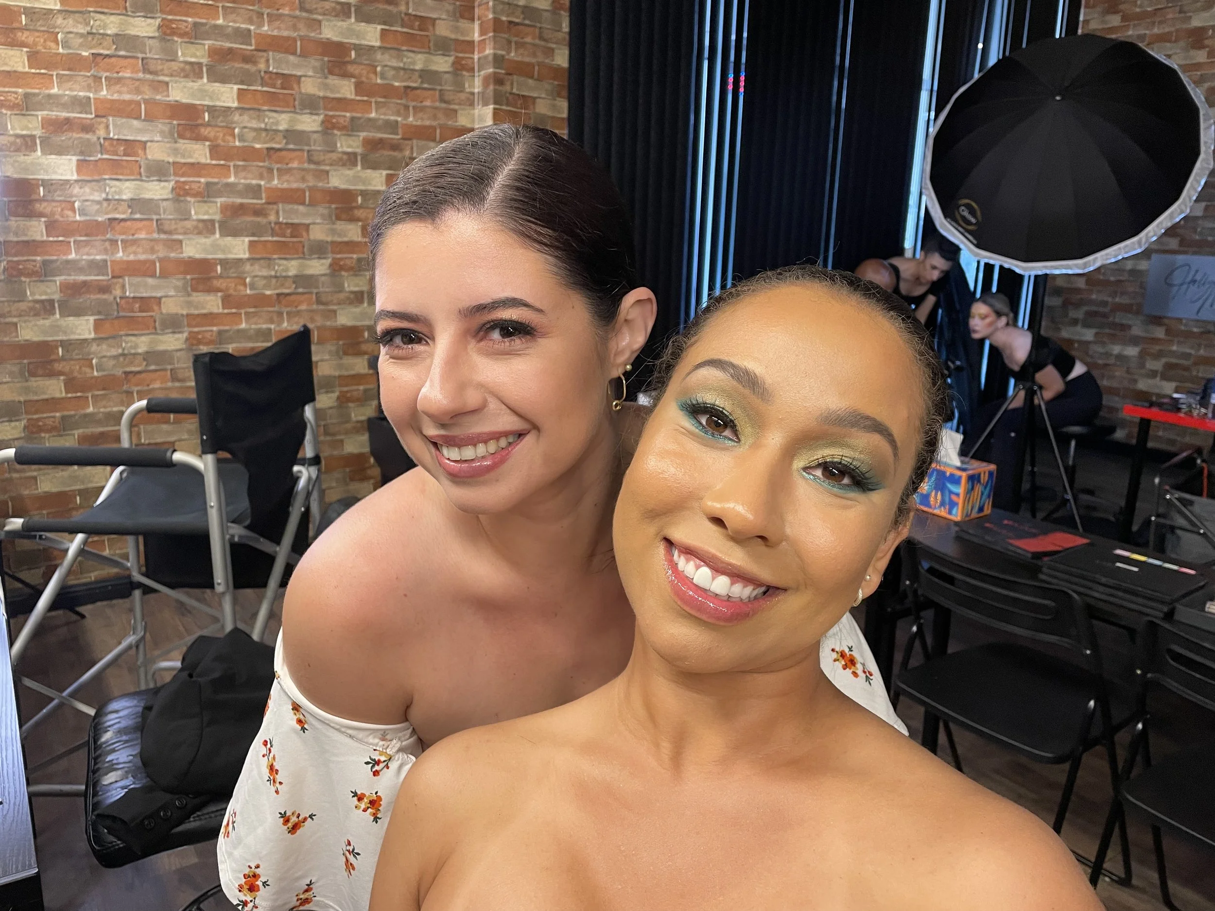 Two women smiling, one with dark hair in an off-shoulder floral dress, the other with curly hair with colorful makeup, in a photography studio with brick wall background and professional lighting equipment.