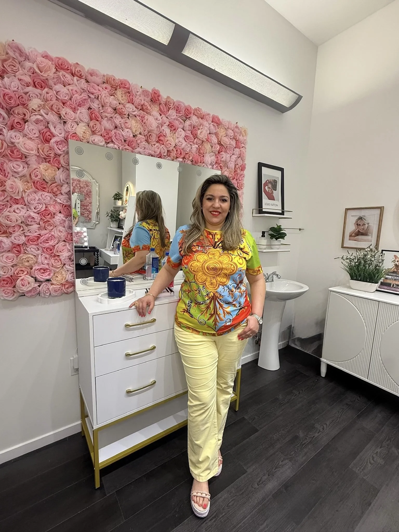 A woman with shoulder-length hair wearing a colorful patterned top and yellow pants, smiling and standing in a room with a wall of pink roses, a mirror, and framed pictures.
