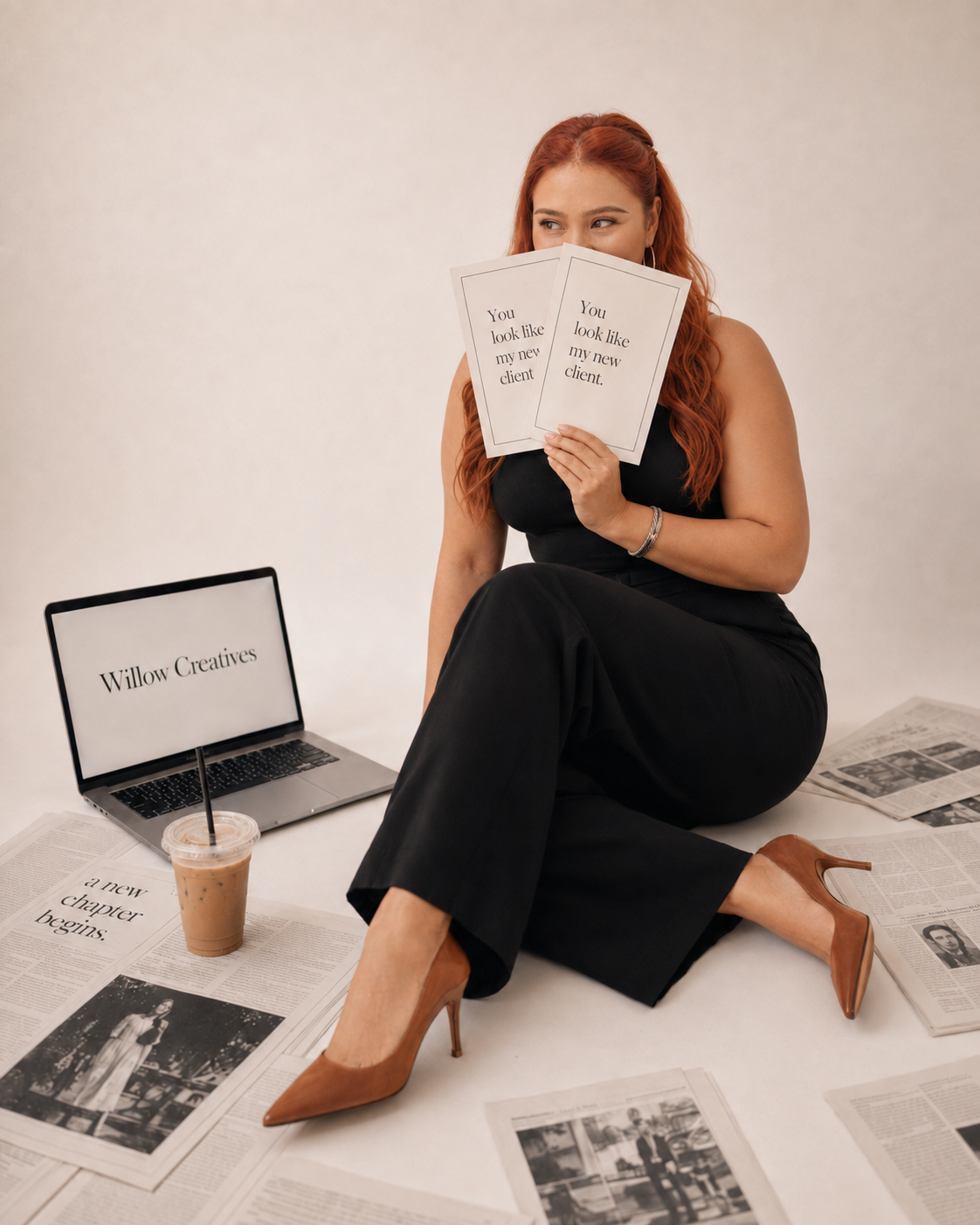 A woman with red hair sitting on the floor among newspapers, holding two sheets of paper with 'You look like my new client' written on them. There is a laptop with the sign 'Willow Creatives' and a cold coffee drink on the floor beside her.