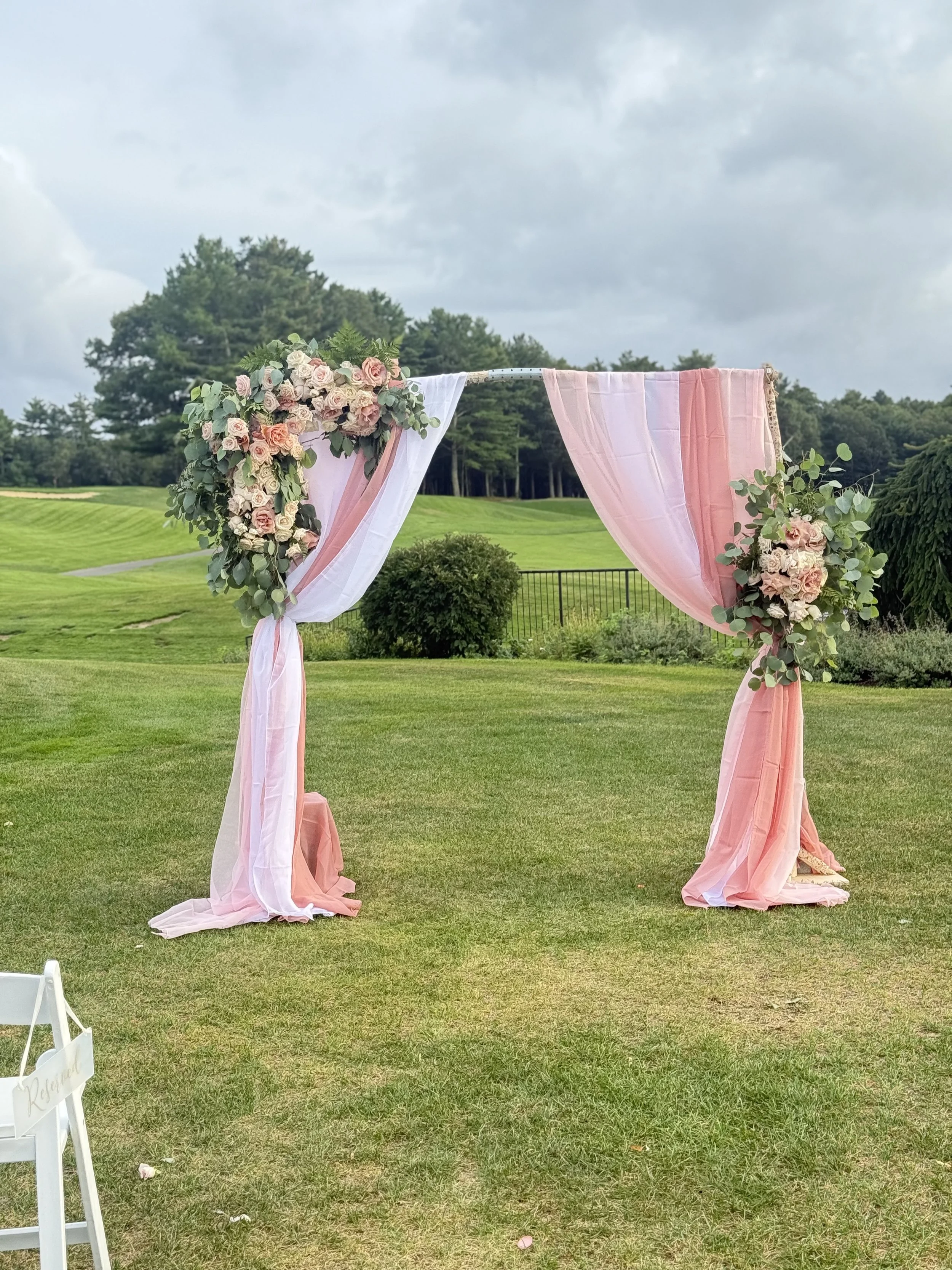 Pink and white draped fabric wedding arbor decorated with flowers, set on a grassy outdoor area with trees and a cloudy sky in the background.