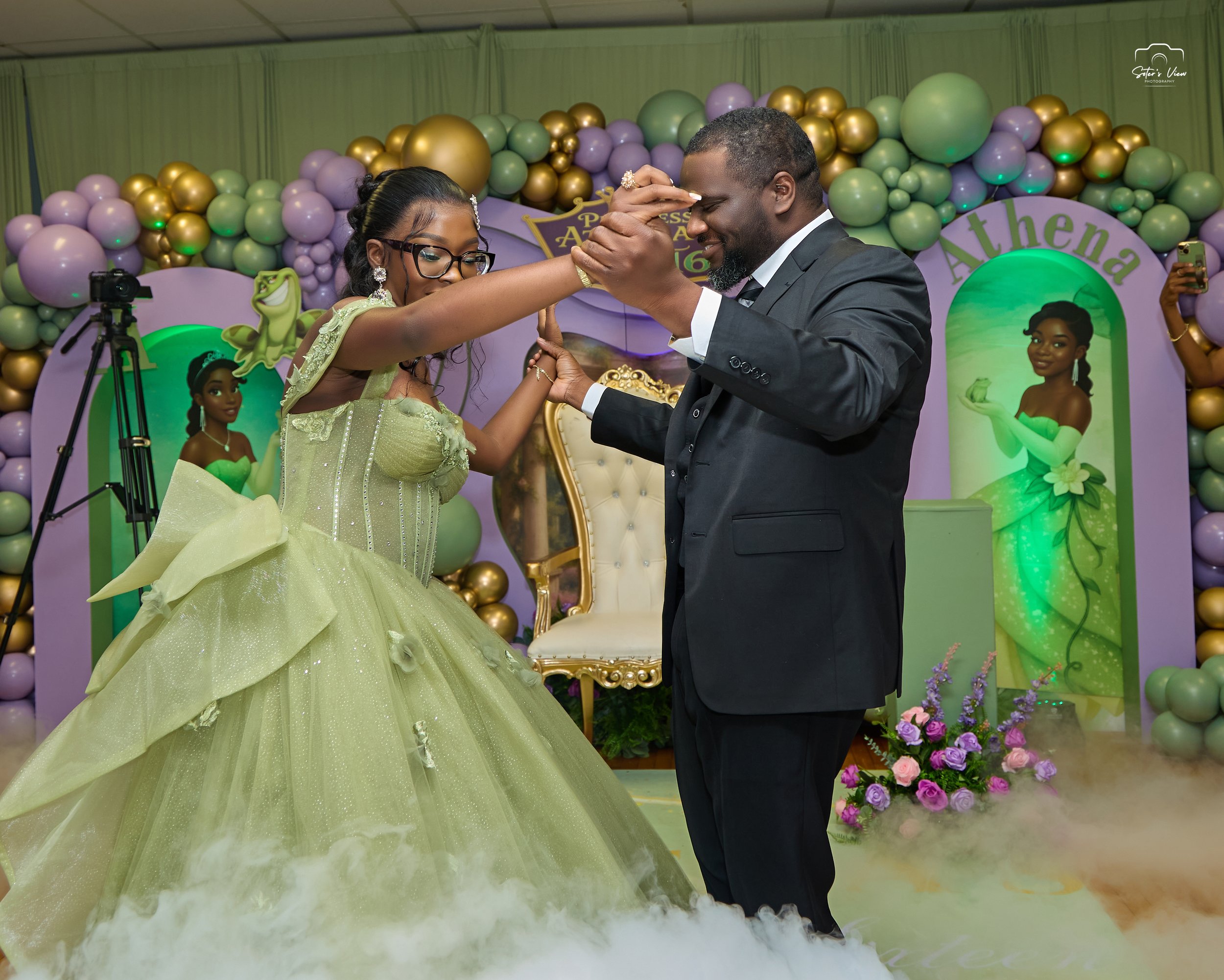 A young woman in a light green gown and glasses dances with a man in a black suit at a celebration event, with a decorated backdrop featuring balloons and a portrait of a woman labeled 'Athena', and floral arrangements.