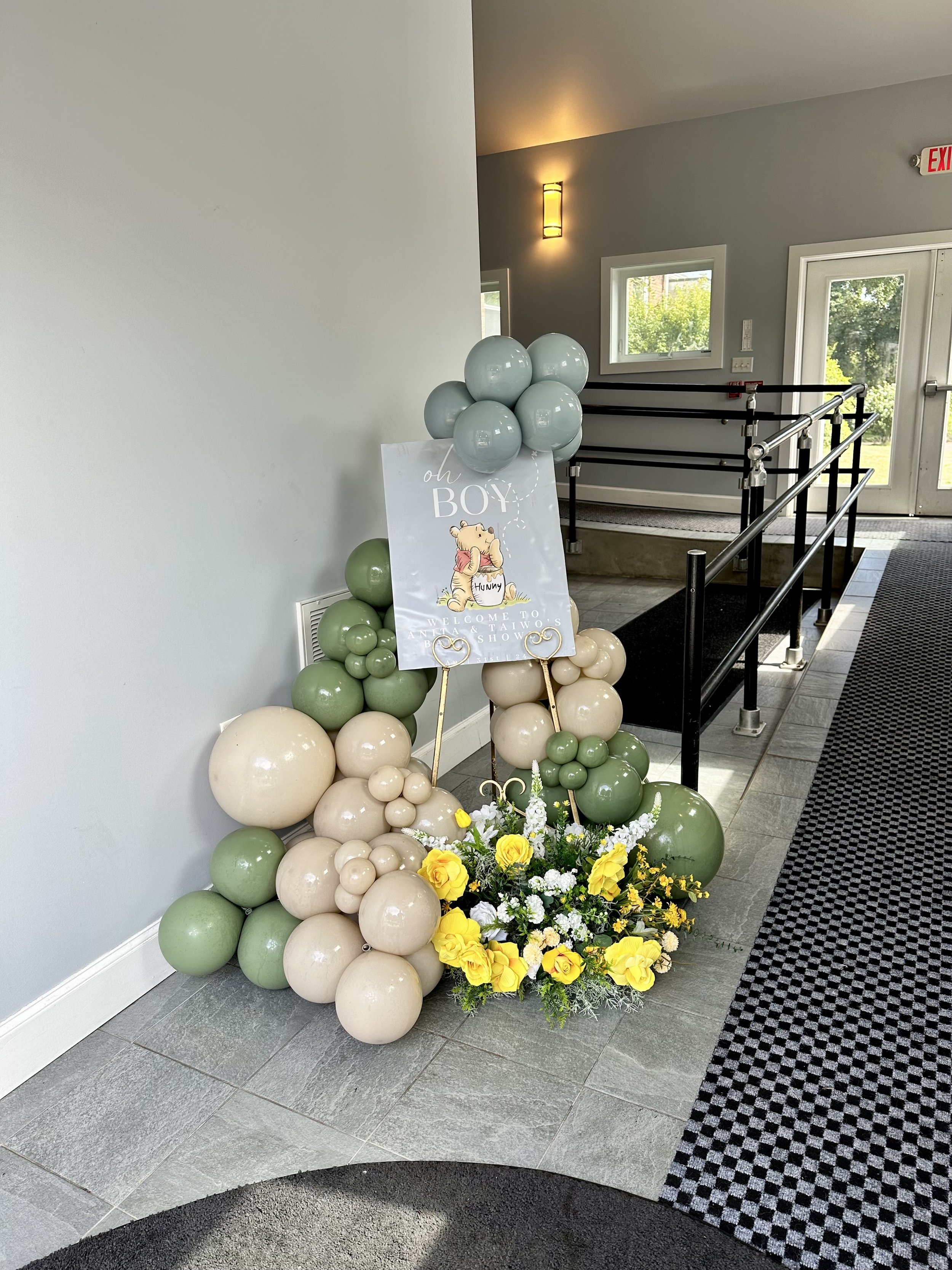 Decorative balloon arrangement with a sign welcoming a newborn boy, featuring cream, light green, and gray balloons, yellow flowers, and a baby-themed sign with a bear illustration.