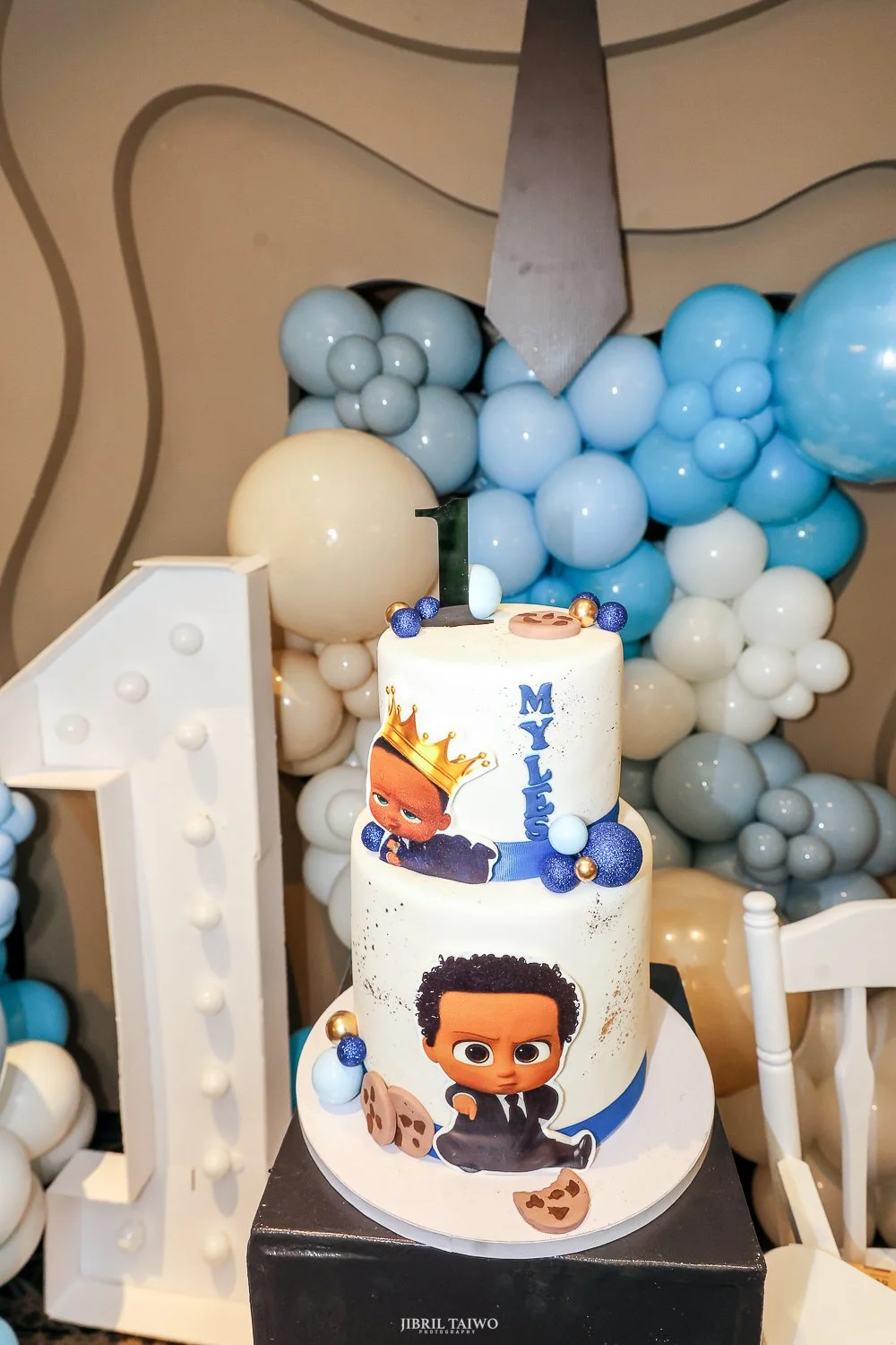 Two-tier birthday cake with cartoon characters of a boy with a crown and a boy with curly hair, decorated with blue, white, and gold balls, with a black '1' candle on top, in front of a balloon backdrop with shades of blue, white, and beige.