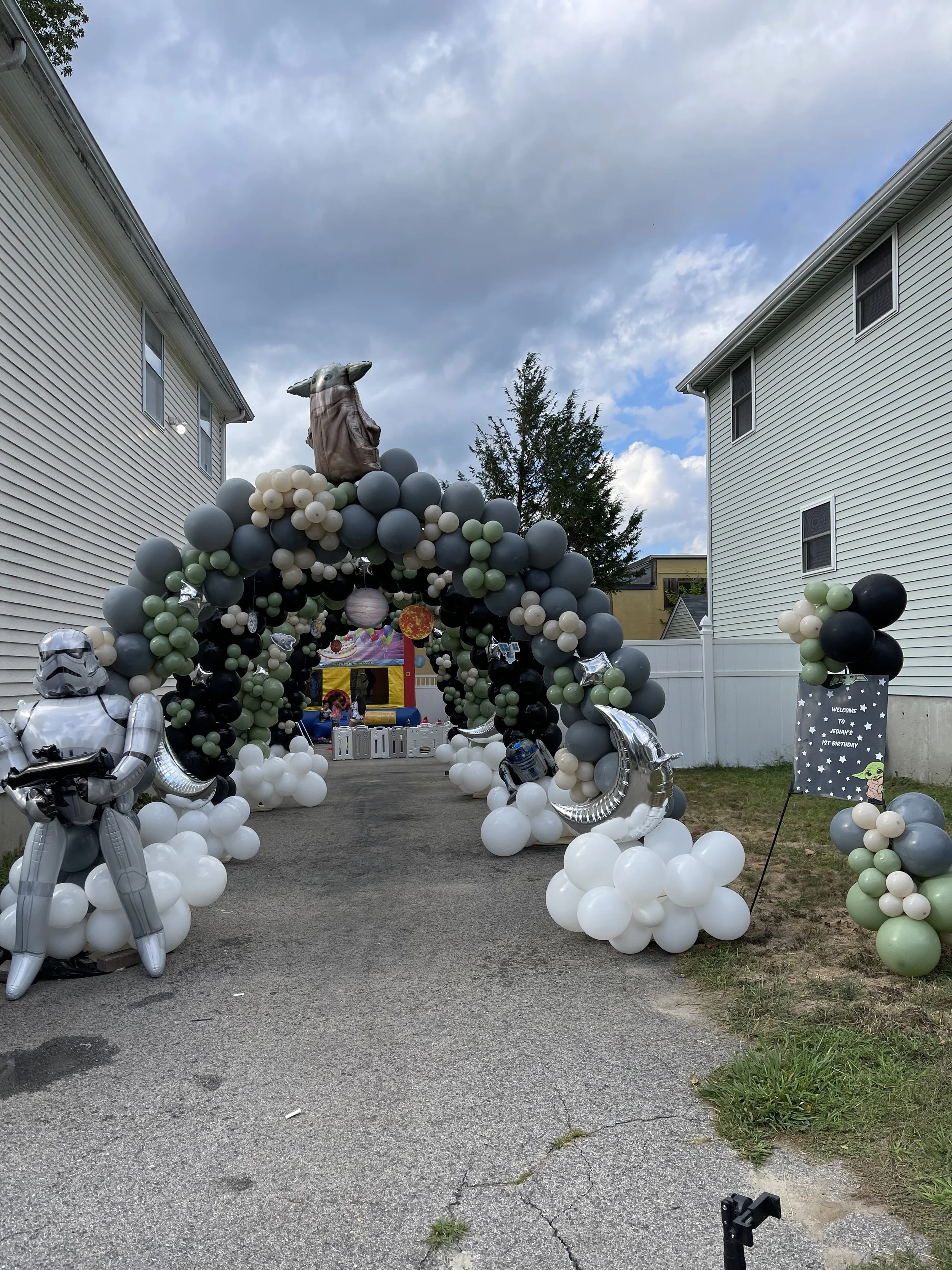 Decorated backyard for a birthday party with a Star Wars theme. There is a balloon arch with black, gray, white, and green balloons, and Star Wars character inflatables, including a Stormtrooper and a Darth Vader. An arch with balloons leads to a bounce house and a sign that reads 'Welcome to Yoda's 1st Birthday'.