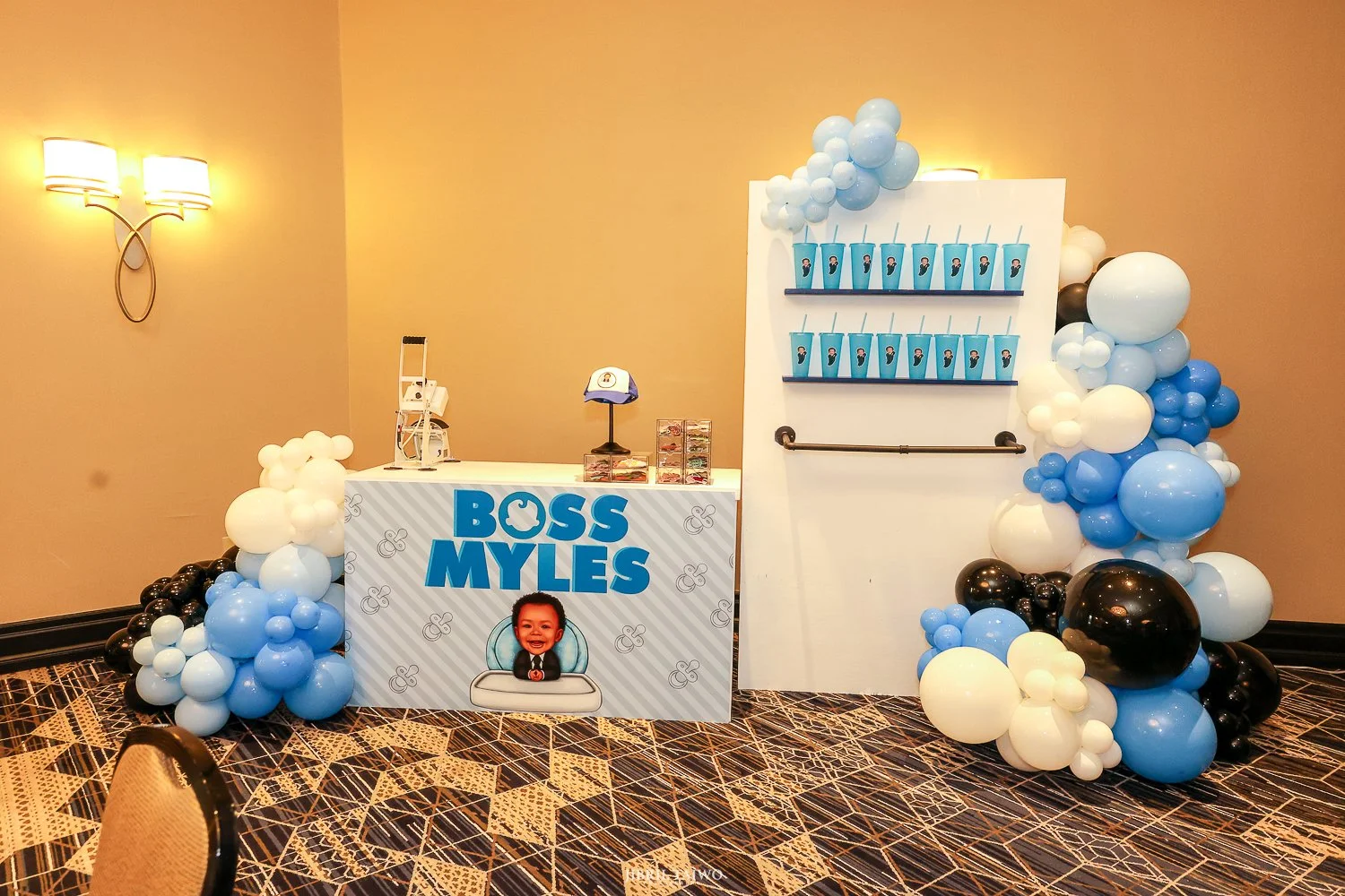 Party display with a sign reading 'Boss Myles' featuring a cartoon of a smiling child in a suit, with blue, white, and black balloons arranged around the display including a table with party favors, and a large white and blue backdrop with drink cups, set in a room with patterned carpet and beige walls.