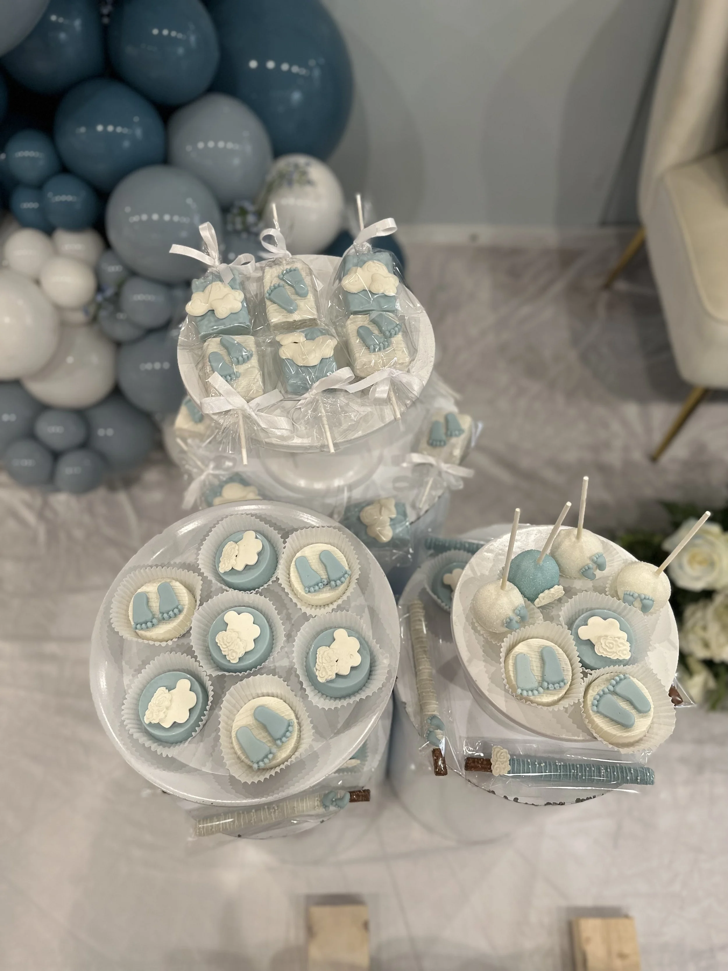 Baby shower table with cupcakes and cake pops decorated with baby-themed blue and white fondant, including baby shoes and sheep motifs, with blue and white balloon decorations in the background.