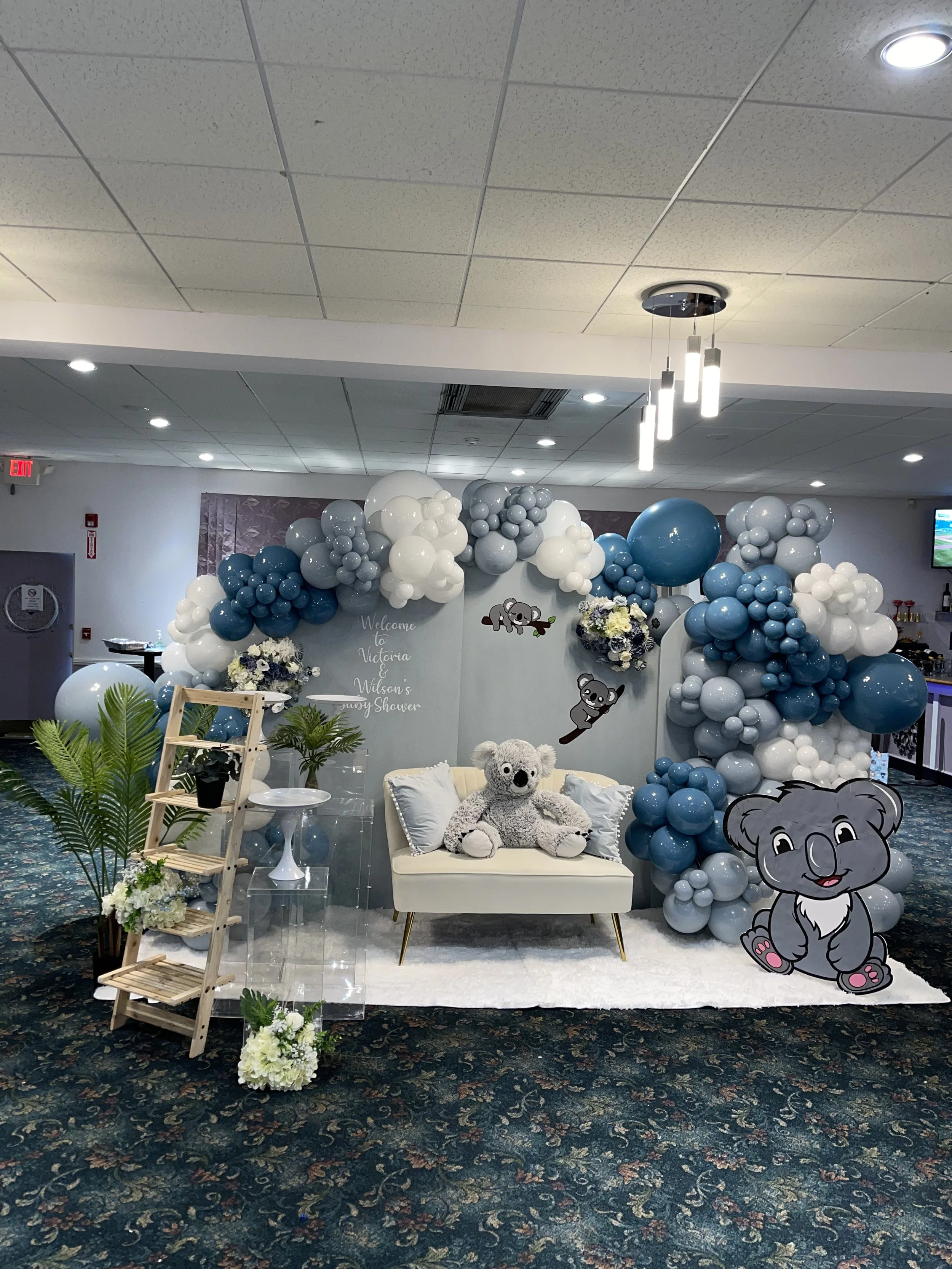 Decorative baby shower setup with a balloon arch in shades of blue, gray, and white, featuring plush koala toys, a small white loveseat, floral arrangements, large cartoon koala cutouts, and a welcome sign.