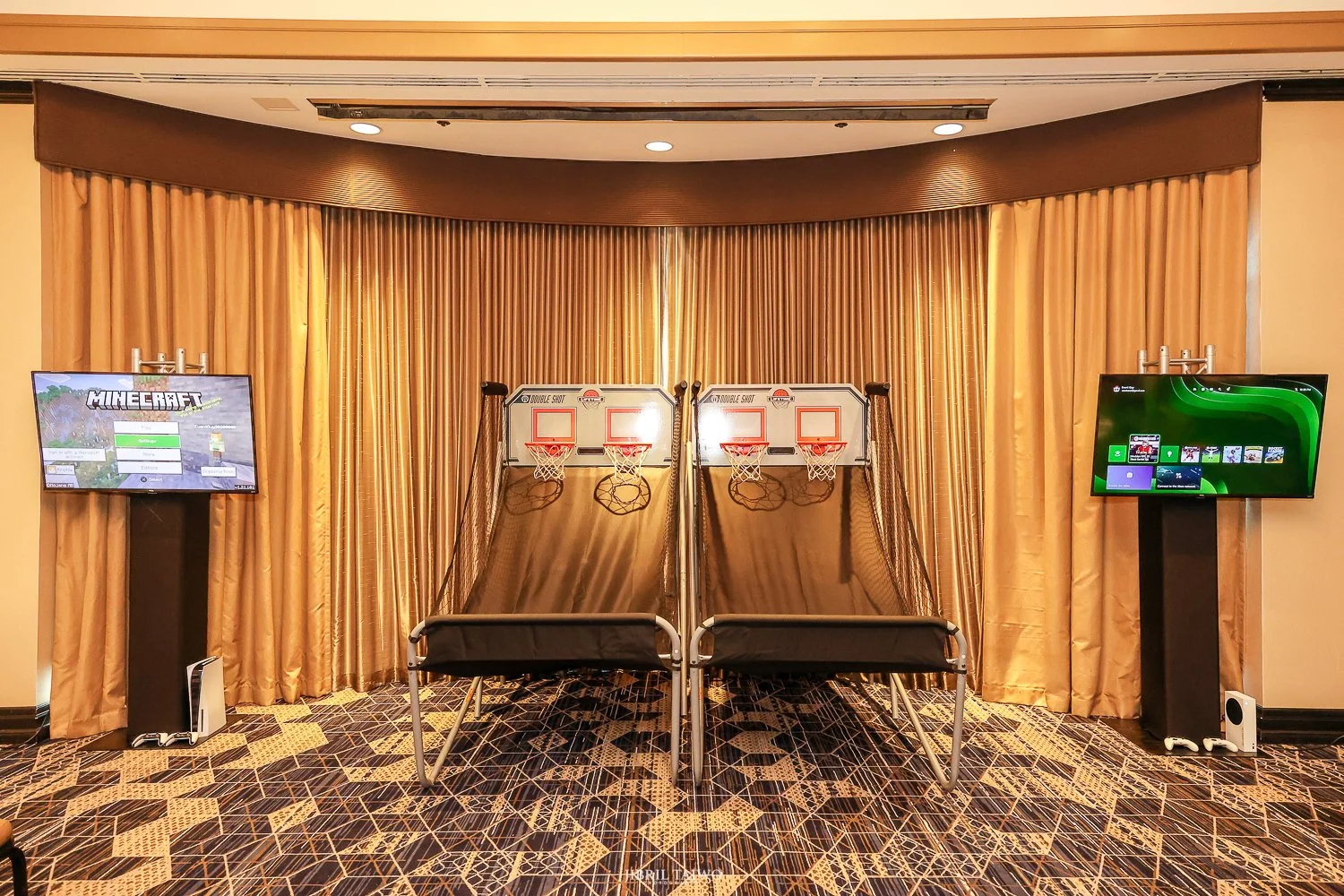 Indoor arcade with two basketball shooting games and two screens on either side, beige curtains in the background, patterned carpet on the floor.