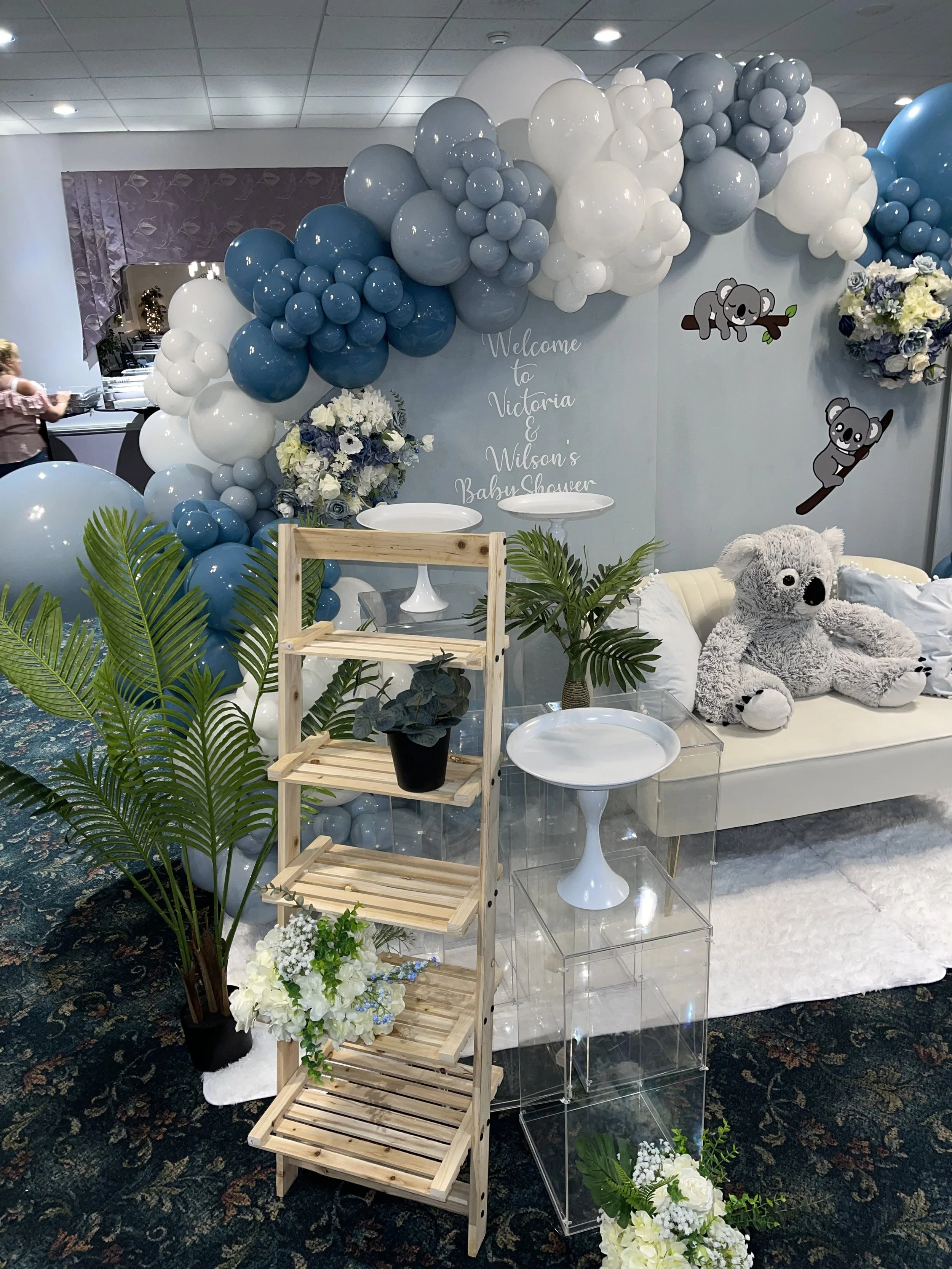 Decorative setup for a baby shower with balloon arch, potted plants, plush koala toy, floral arrangements, and a white couch.