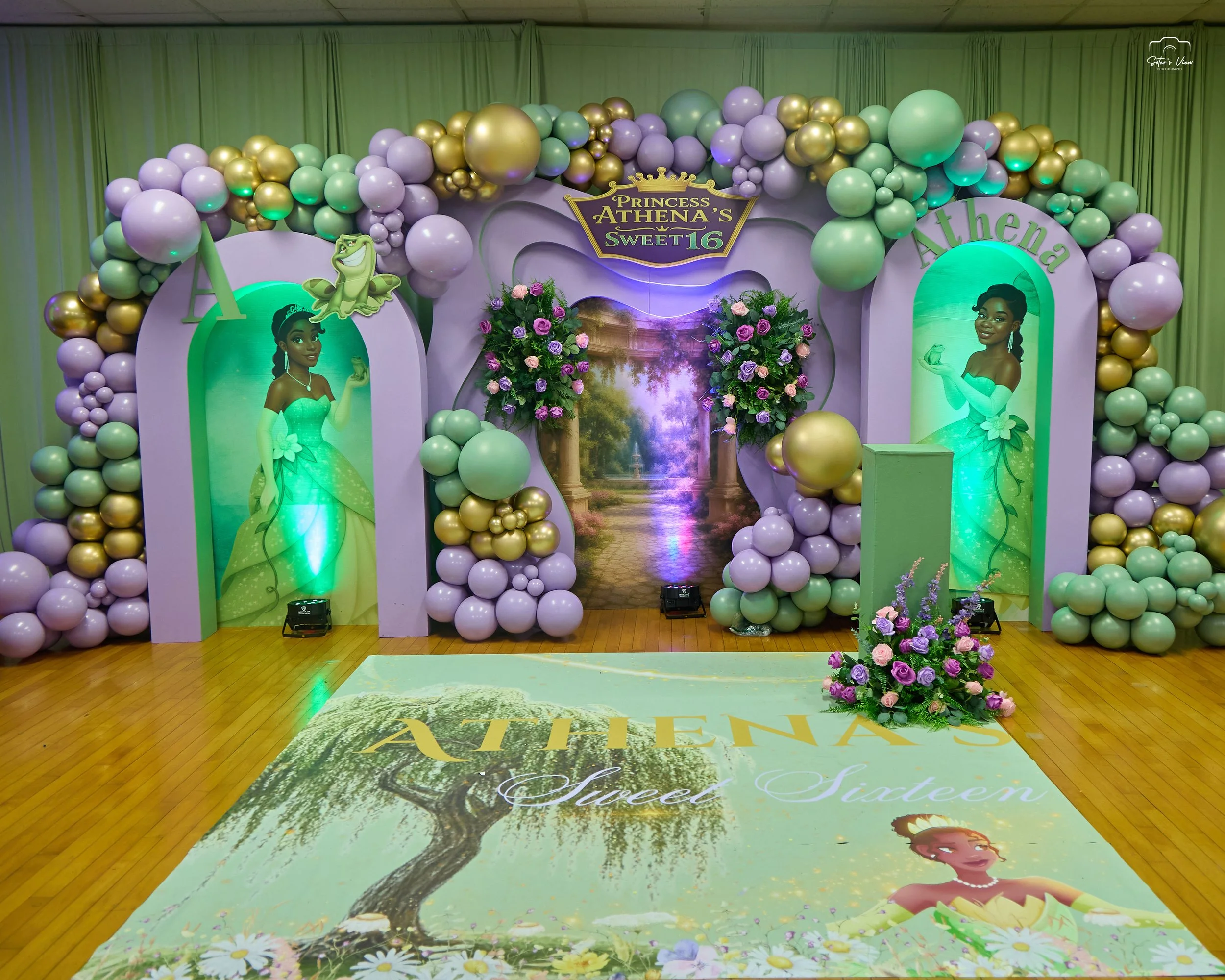 Decorative setup for a princess themed celebration with balloons in lavender, gold, and mint green, floral arrangements, and a backdrop with a pathway and nature scene. The floor has a printed design reading "Athena's Sweet Sixteen" with a princess illustration.