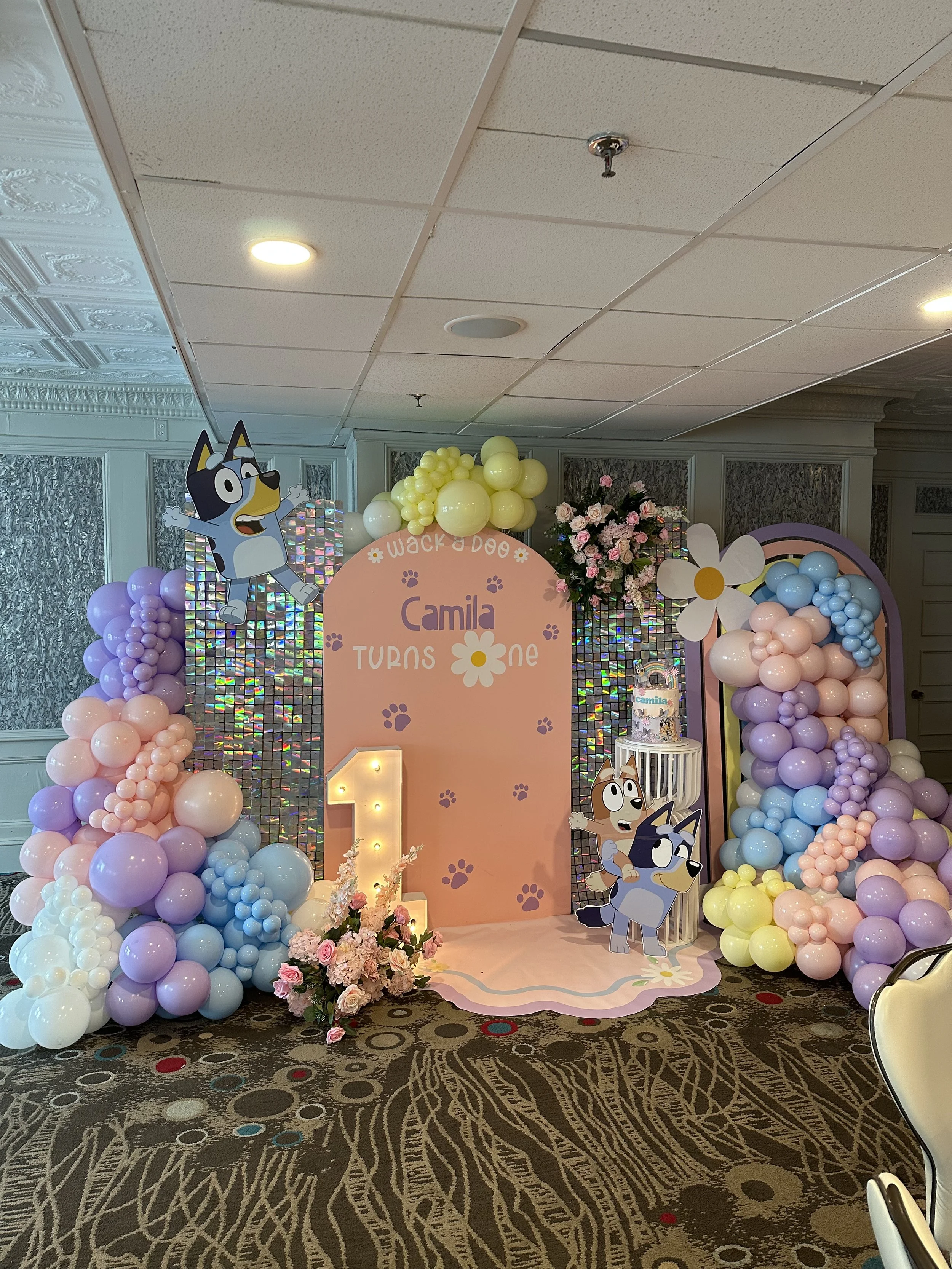 Decorative party backdrop with balloon arrangements in pastel colors, a large number one, cartoon dog characters from Paw Patrol, and a cake with a rabbit figure, for a child's first birthday celebration.