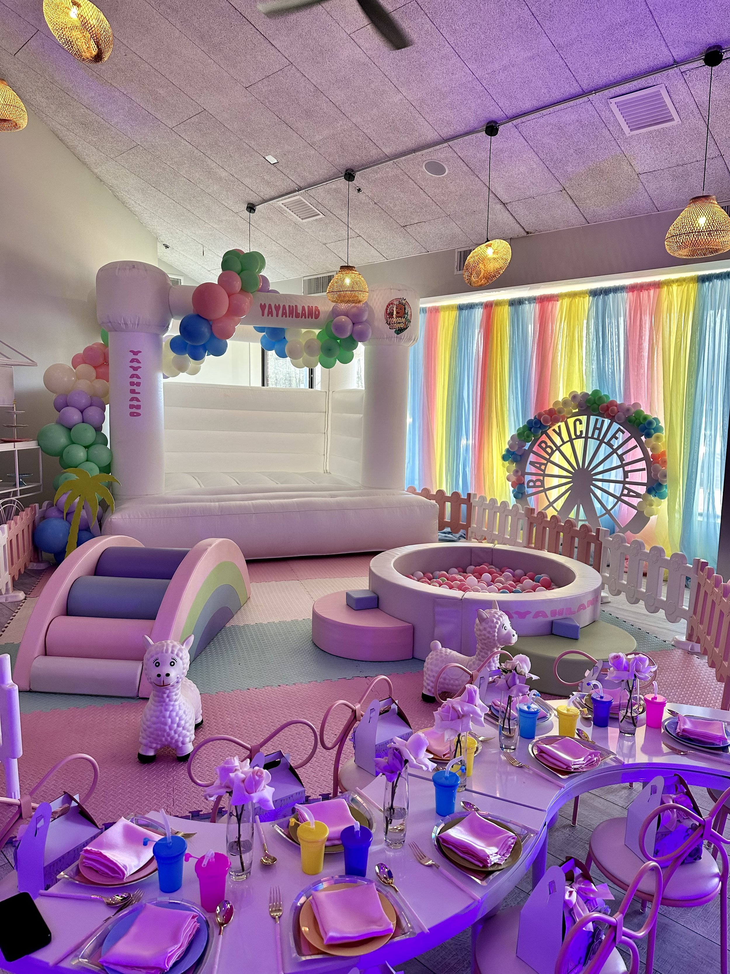 Colorful birthday party room with pastel decorations, a ball pit, a rainbow slide, balloons, a Ferris wheel, and a table set with plates, napkins, and drinks for children.