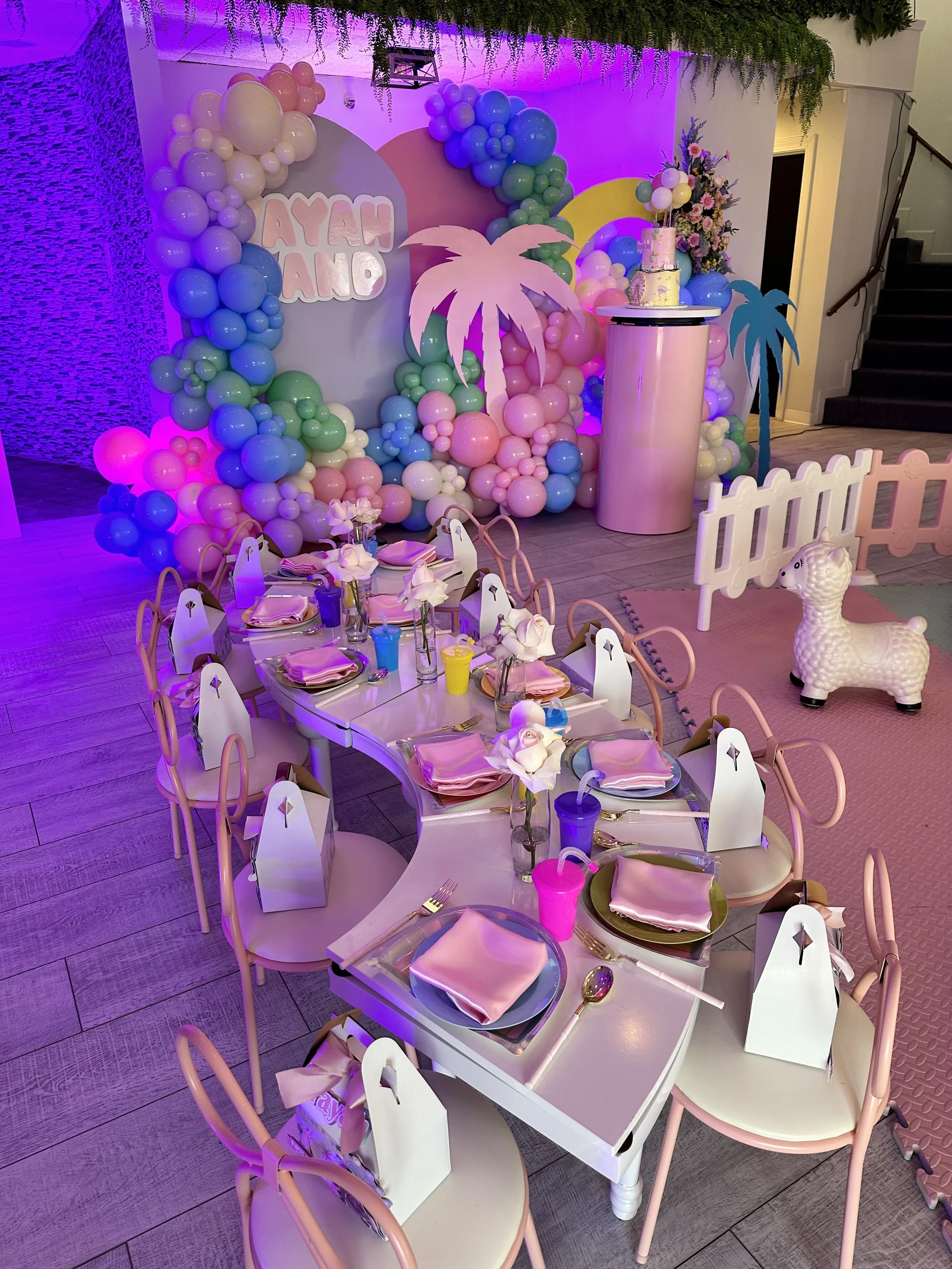 Decorated party setup with pastel balloons, a table set with pink napkins and flowers, and a pink and purple theme with a cake and inflatable toys.