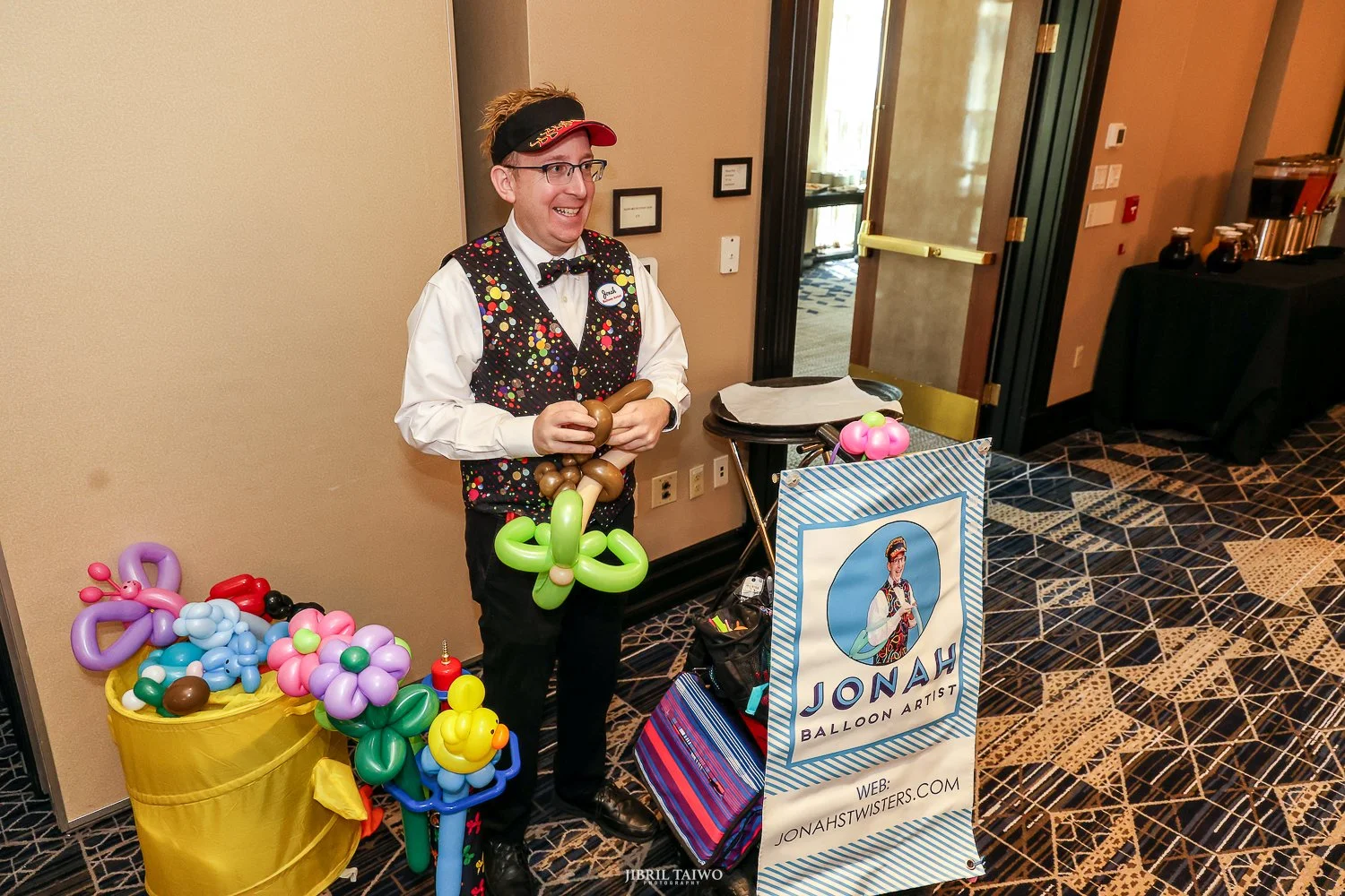 A balloon artist dressed in a colorful vest, bow tie, and glasses standing at a booth with balloon sculptures, smiling at an event.