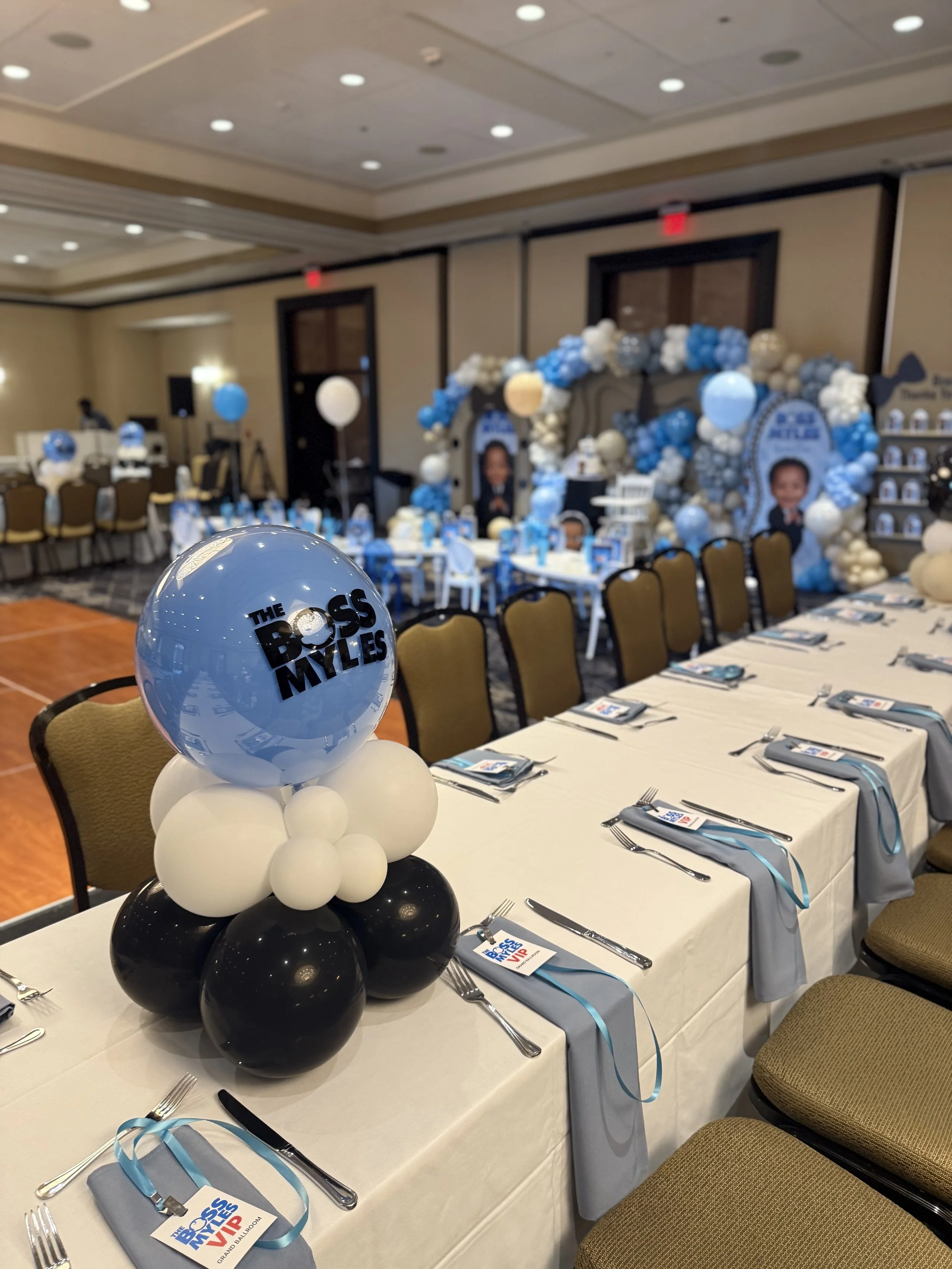 Event space decorated in blue, white, and black theme for a celebration, with balloon arrangements, tables set with utensils and ribbons, and a large backdrop with balloons and photos, celebrating a child named Miles with the theme "The Boss Miles."