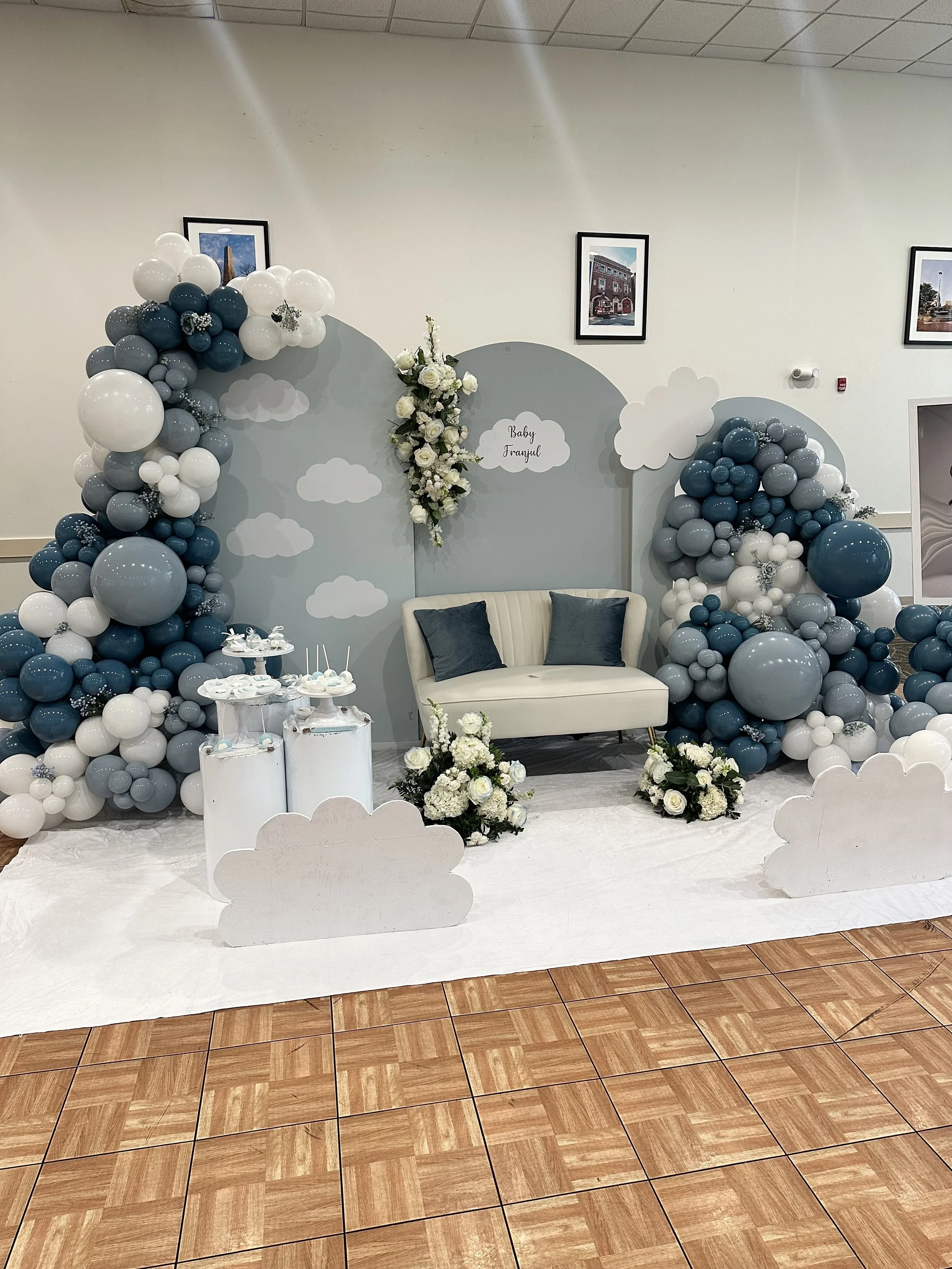 Decorated backdrop for a baby shower with clouds and balloon arrangements in shades of blue, white, and gray, featuring a small sofa, floral arrangements, and cloud cutouts marking it as a 'Baby Shower' celebration.