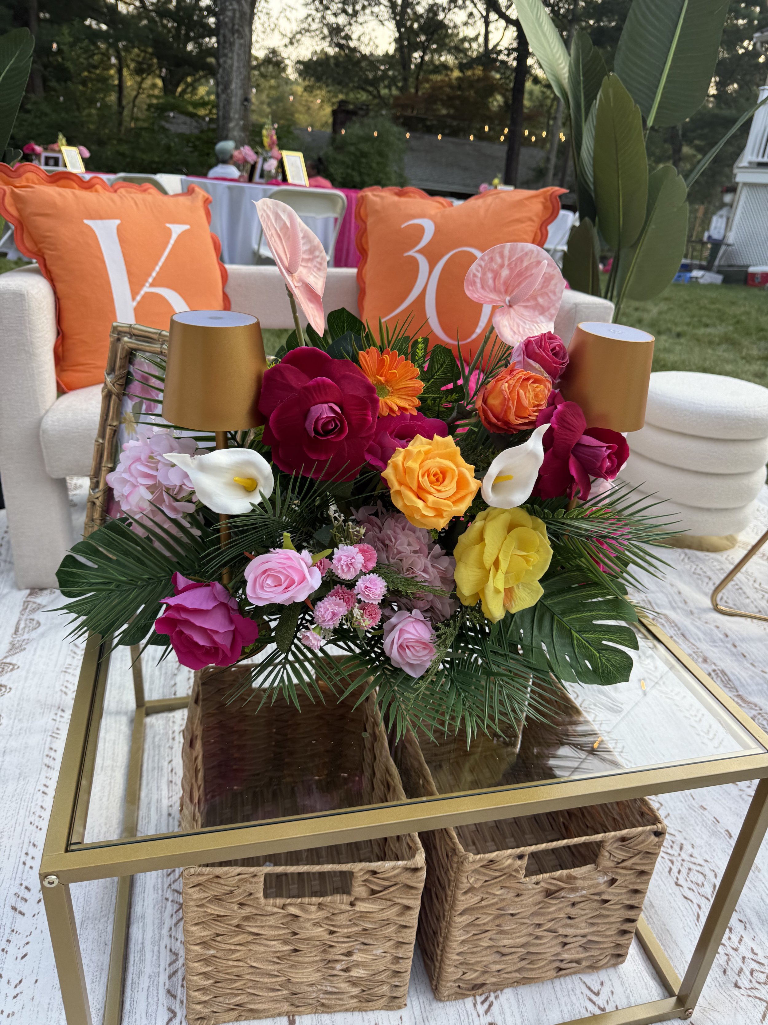 A floral centerpiece on a glass-top table with pink, yellow, red, and orange flowers, two small gold lamps, and two wicker baskets underneath. In the background, there are orange pillows with white letters and a white sofa with additional décor.