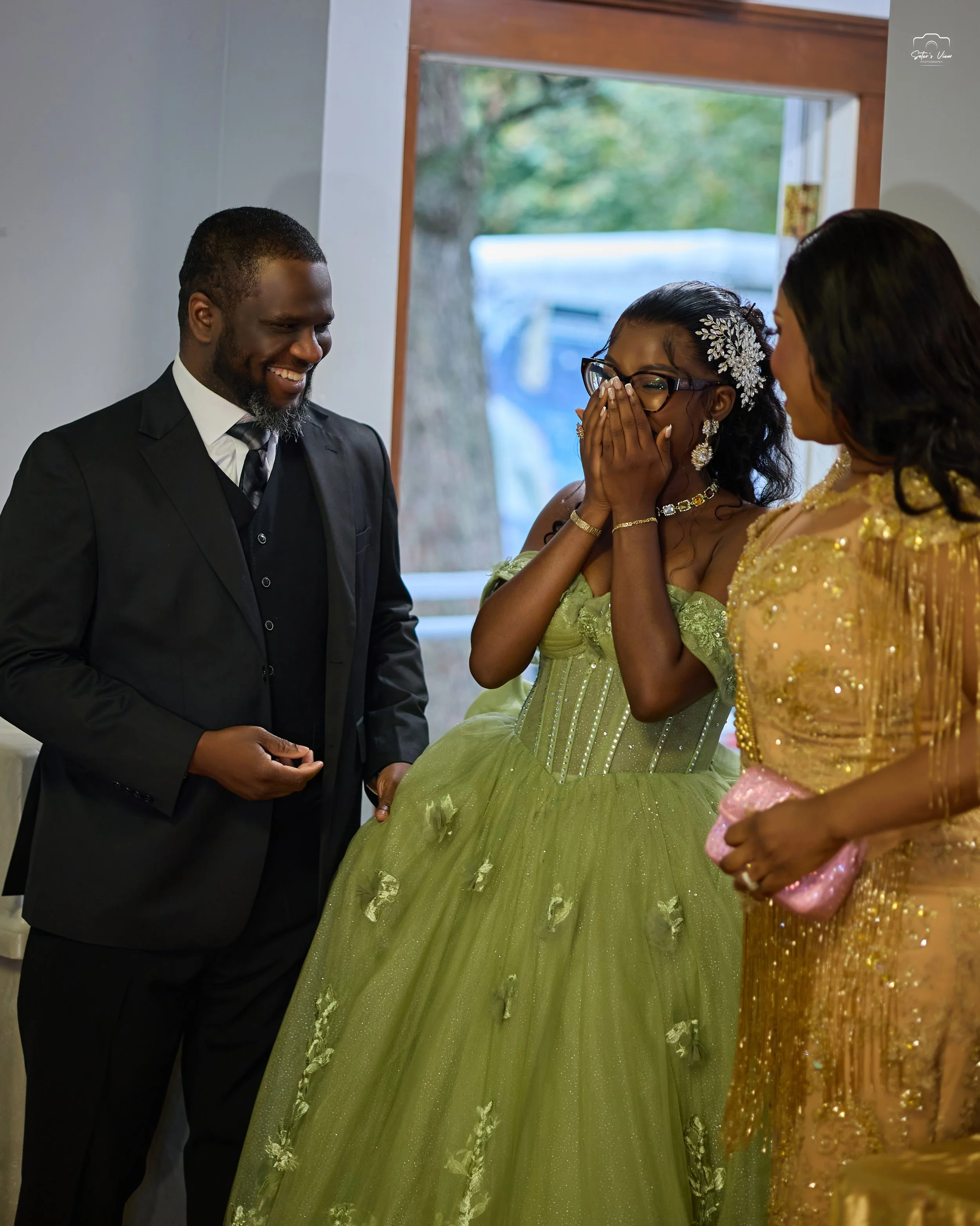 A groom in a black suit smiling at a bride dressed in a green gown with an emotional expression, while a woman in a gold gown looks on, in an indoor setting.