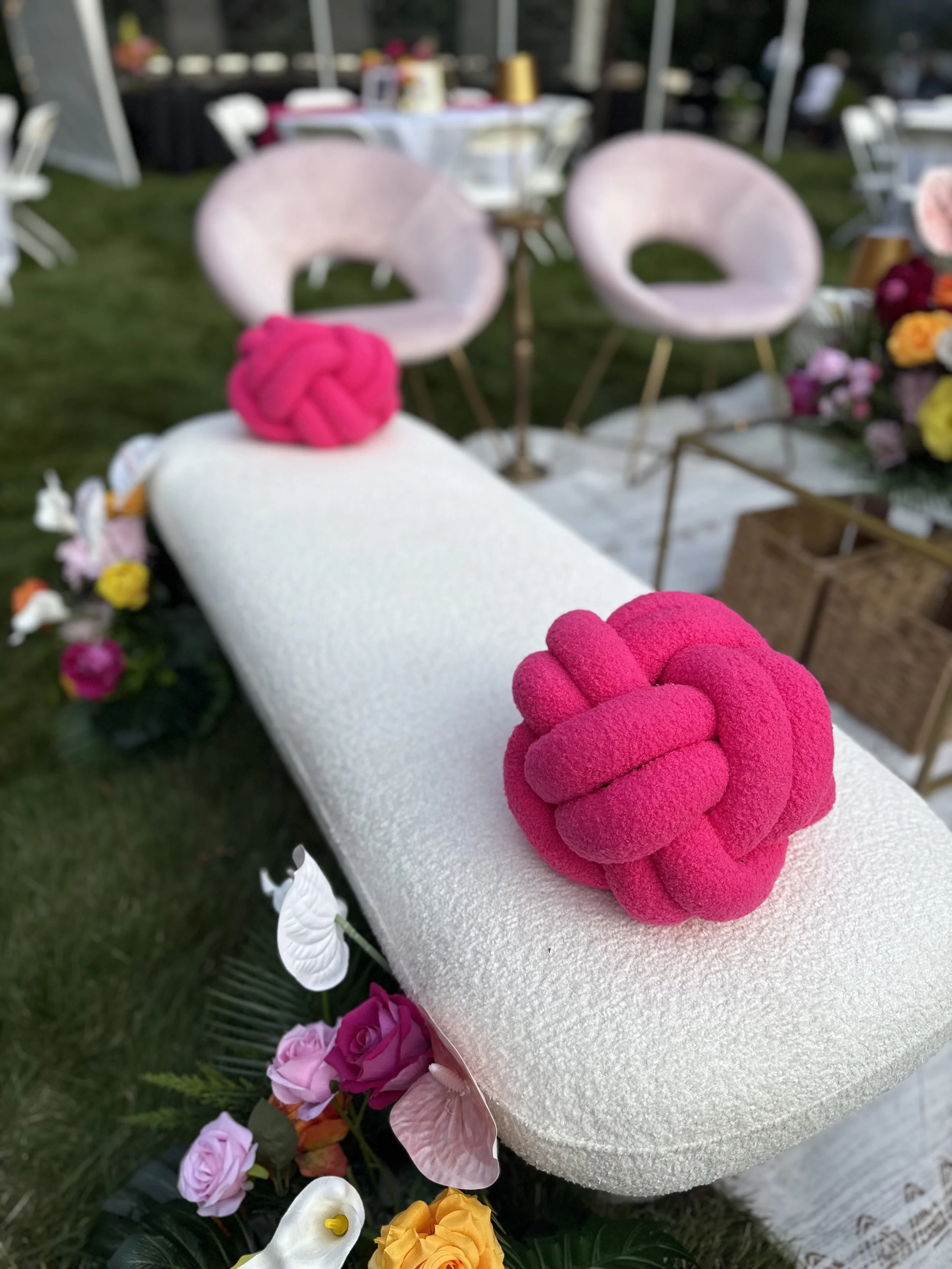 Decorative wedding or event setup with a white textured platform adorned with pink knotted plush cushions, surrounded by flowers, including roses and anthuriums, and with pink velvet chairs in the background.