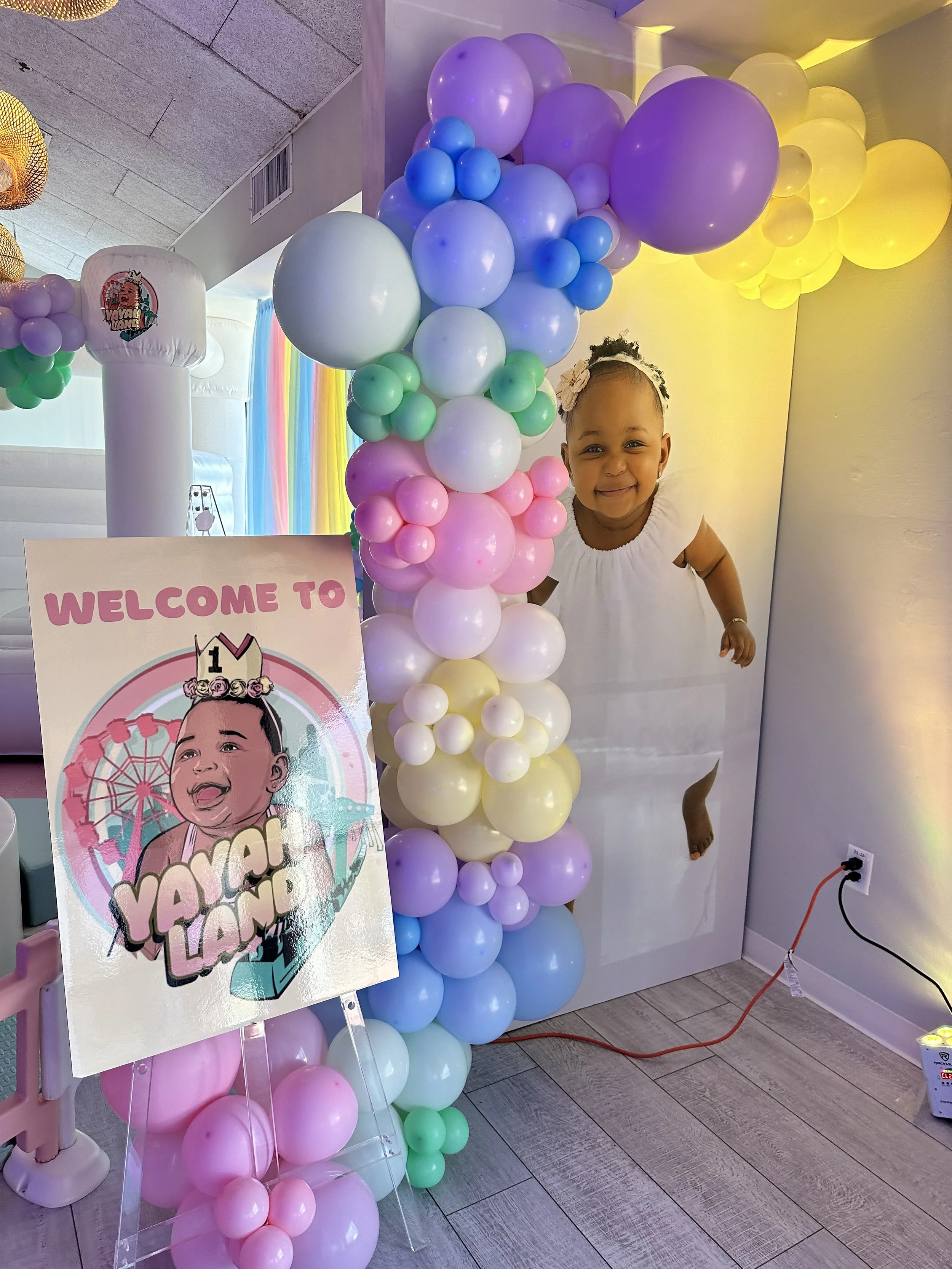 A young girl with a big smile standing behind colorful balloon decorations and a birthday sign celebrating a first birthday with the theme 'Yay for Lana'.