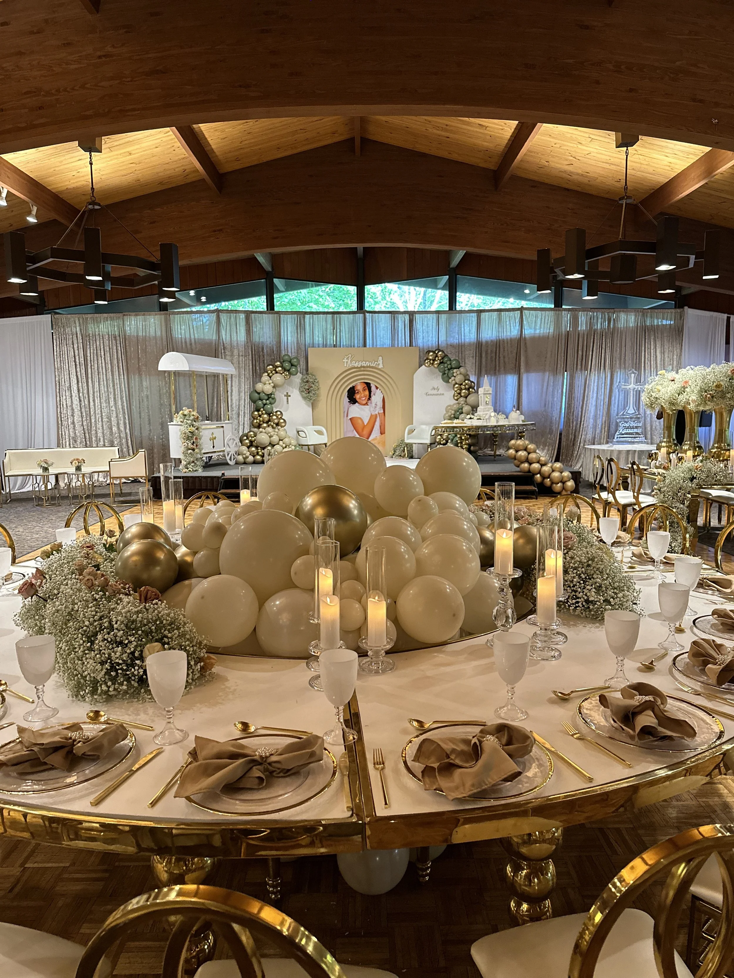 Elegant banquet hall decorated for a celebration with a centerpiece of white and gold balloons, surrounded by candles, flowers, and set tables with gold accents.