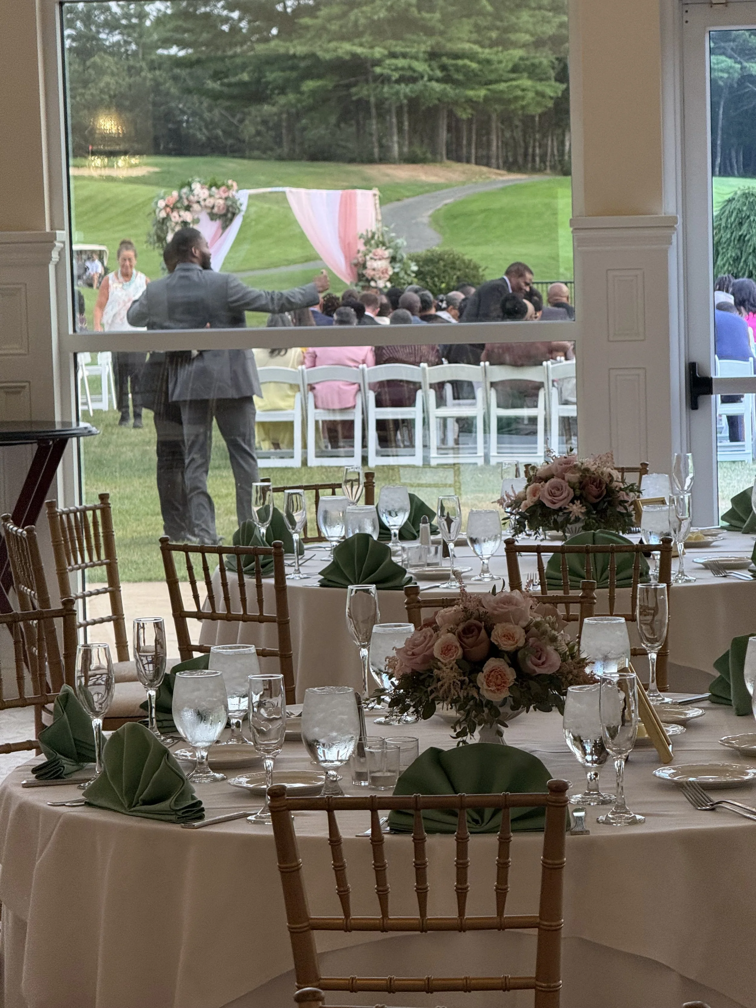 Indoor wedding reception area with decorated tables, flowers, glassware, and green napkins, viewing an outdoor wedding ceremony with guests, chairs, floral arrangements, and a pink and white draped arch on a grassy lawn.